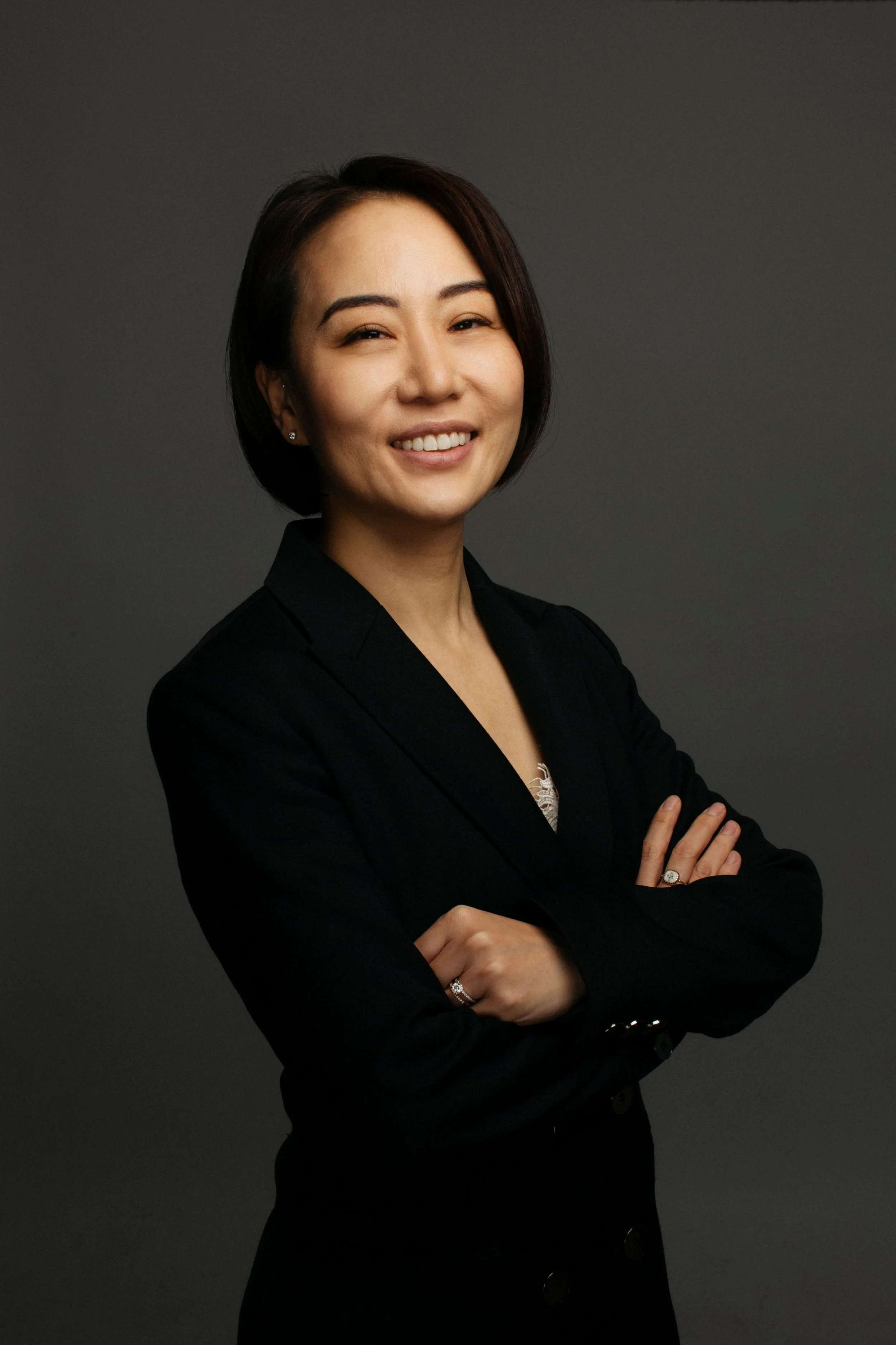 A professional woman smiling with crossed arms, wearing a black blazer and earrings, against a dark gray background.