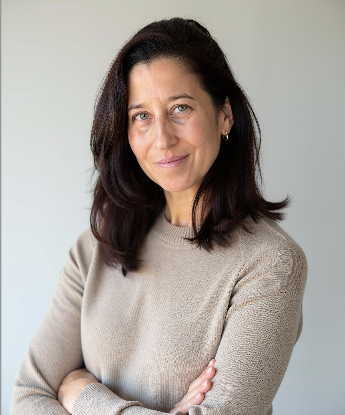 A woman with shoulder-length dark brown hair and light eyes, wearing a beige long-sleeve top, standing against a plain light gray background, looking slightly to her right with a soft smile.