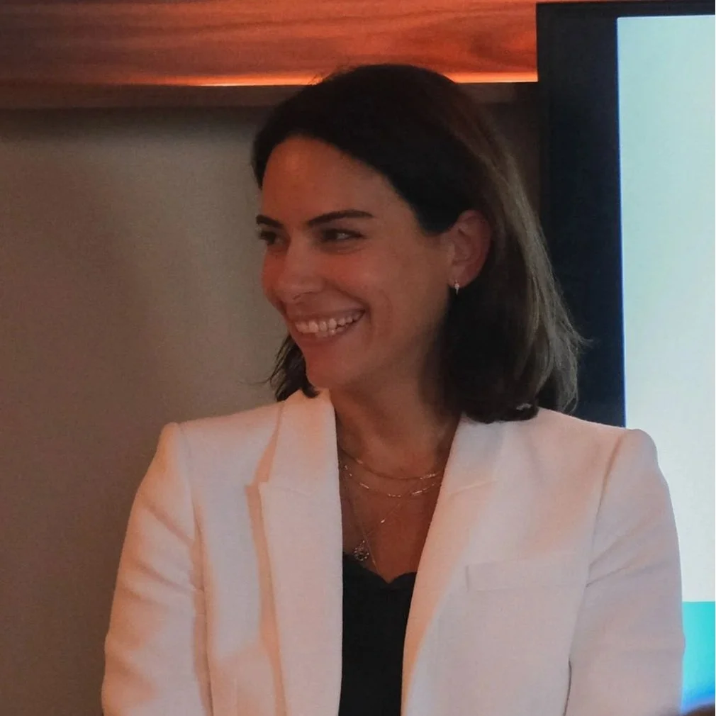 A woman in a white blazer smiling and looking to her left indoors.