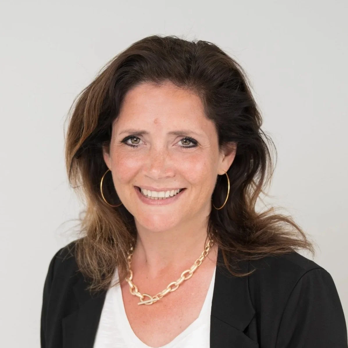 A woman with mid-length brown hair, wearing a black blazer, white top, gold hoop earrings, and a gold chain necklace, smiling against a plain white background.