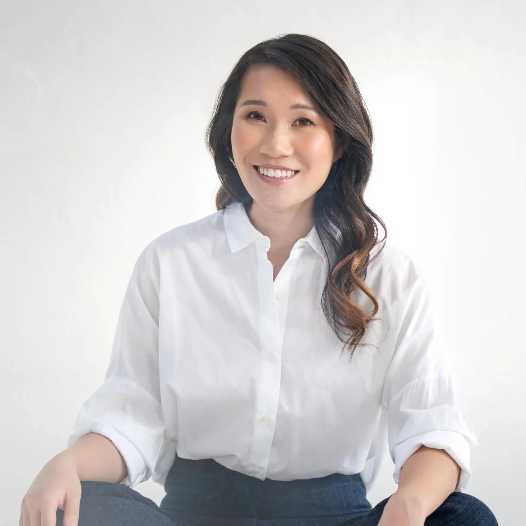 A woman with long dark wavy hair, smiling, wearing a white button-up shirt with rolled-up sleeves and dark jeans, sitting against a plain white background.