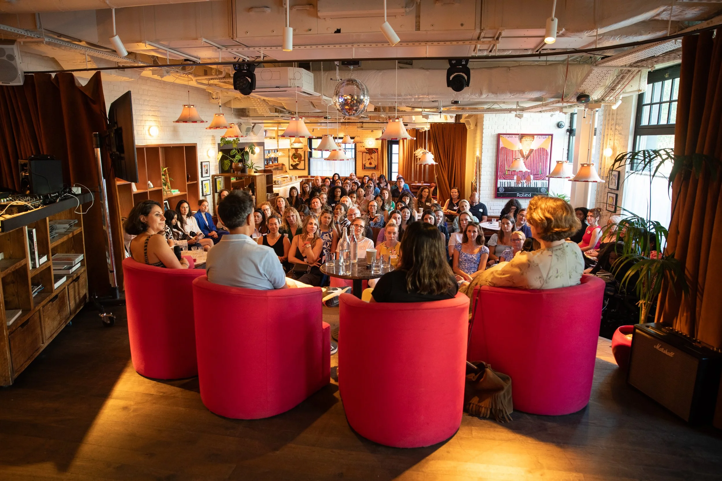 Audience seated facing a speaker, with a woman in a black dress speaking, in a room with warm lighting, wooden furniture, plants, and entertainment posters.