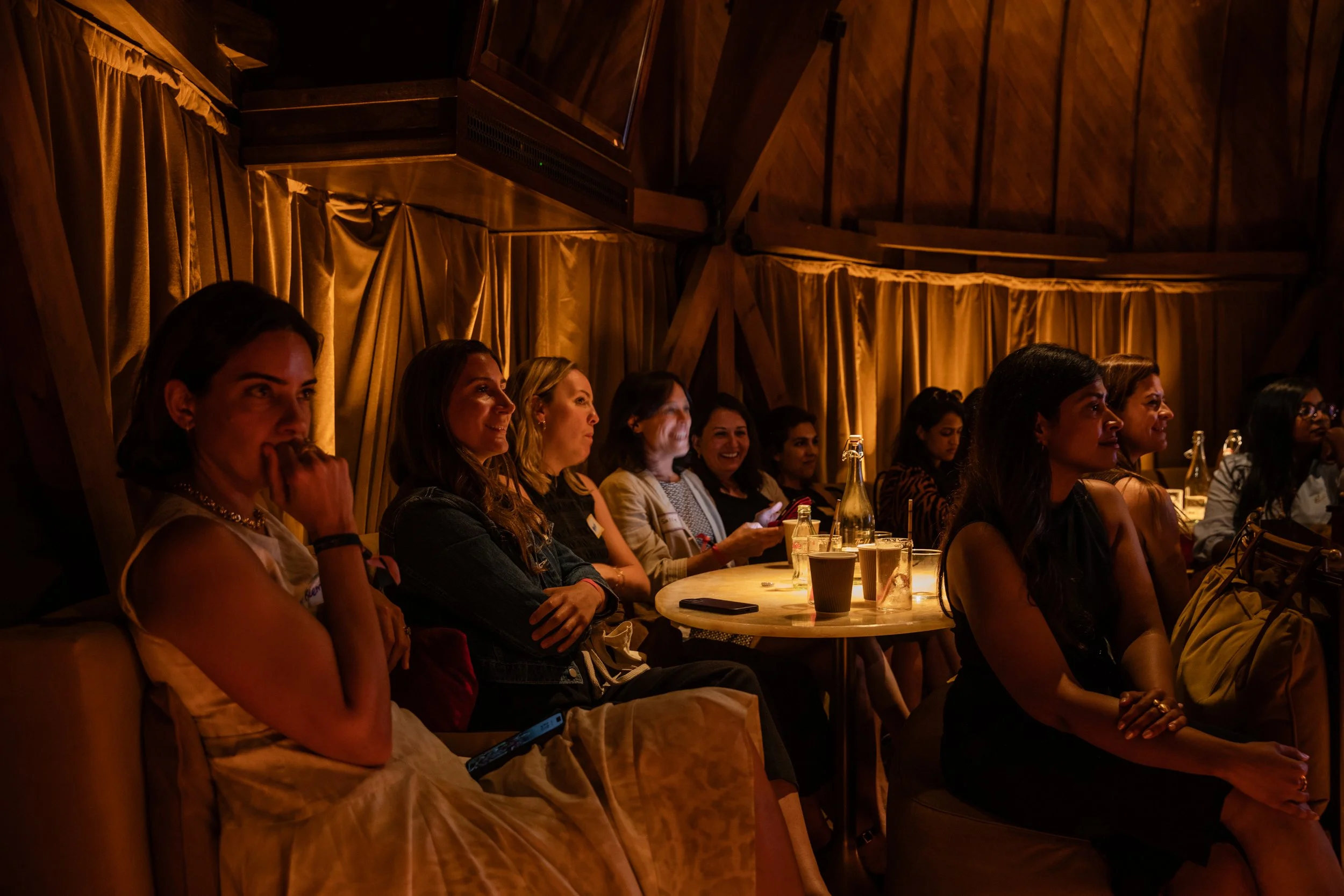 A group of women sitting at a round table in a dimly lit venue, watching a performance or speaker. The setting has wooden walls, golden curtains, and drinks on the table.