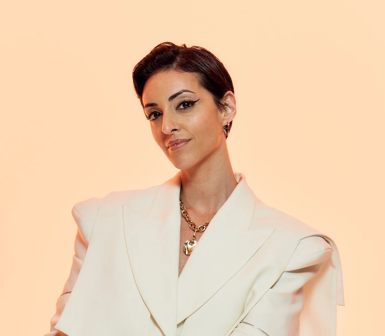 A woman with short dark hair wearing a white blazer and gold jewelry, posing against a peach-colored background.