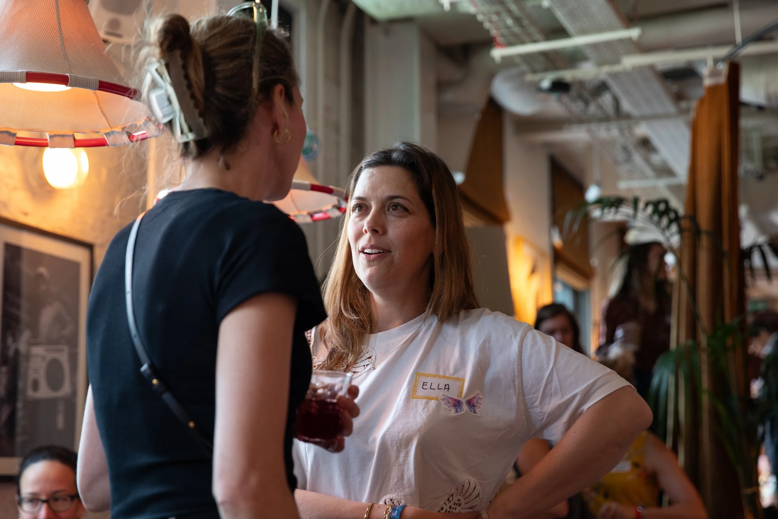 Two women are engaged in a conversation in a busy indoor setting. One woman is wearing a black shirt with a name tag that says "ELLEN," and the other woman has long brown hair and is wearing a white shirt. The background shows other people, plants, and hanging lights, suggesting a social or family event.