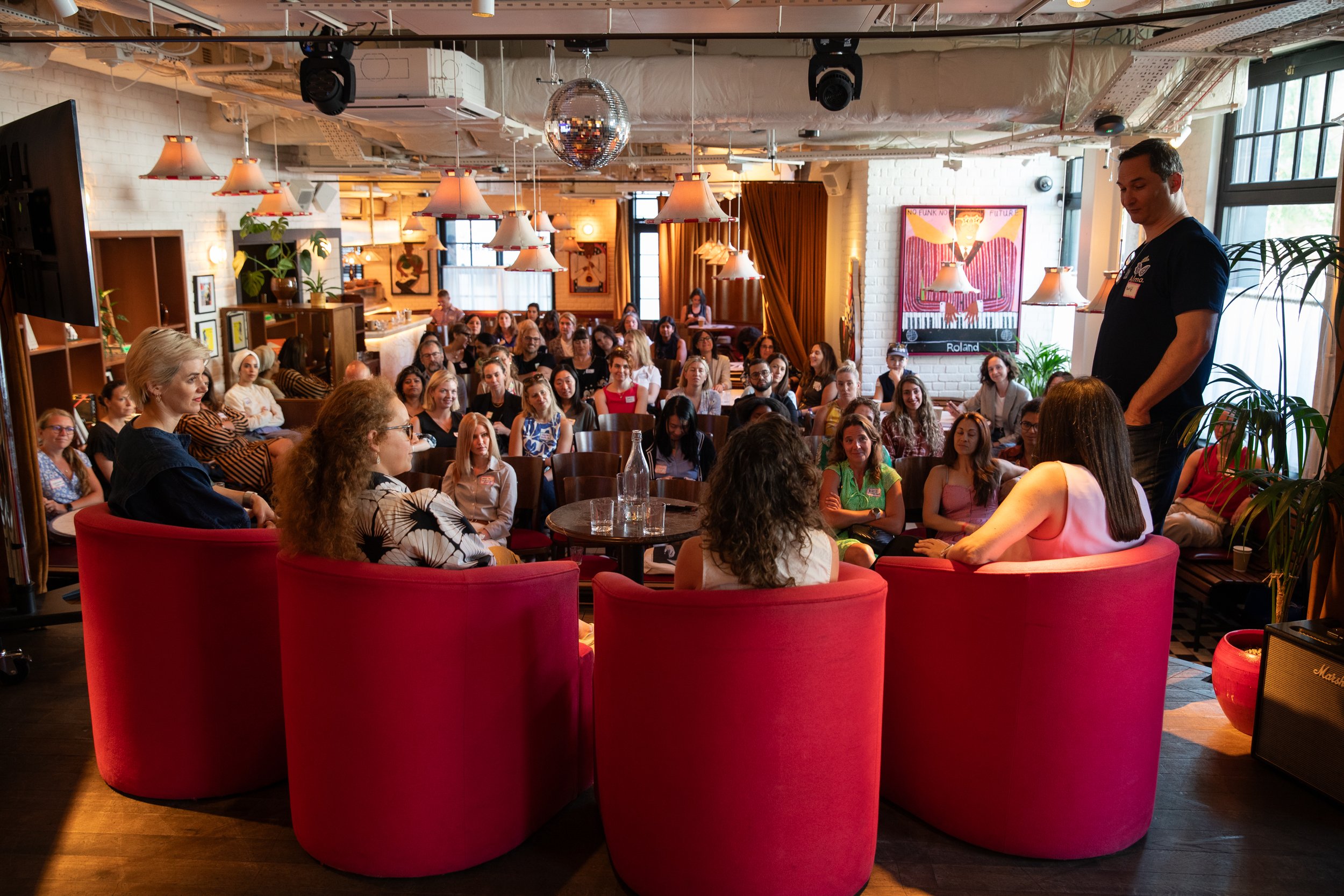 A large gathering of people seated in a cozy, well-lit cafe or restaurant with modern decor, including hanging lamps, potted plants, and artwork on the walls. A man stands in front of the audience, possibly speaking or presenting.