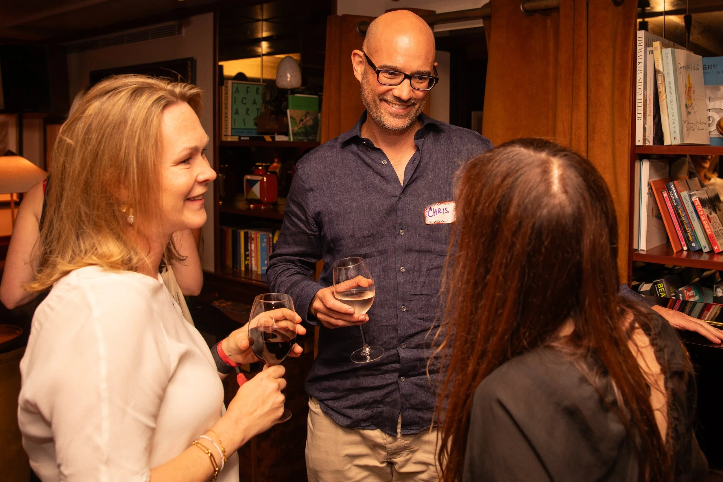 People socializing at a gathering, holding wine glasses, in a cozy indoor setting with bookshelves and warm lighting.