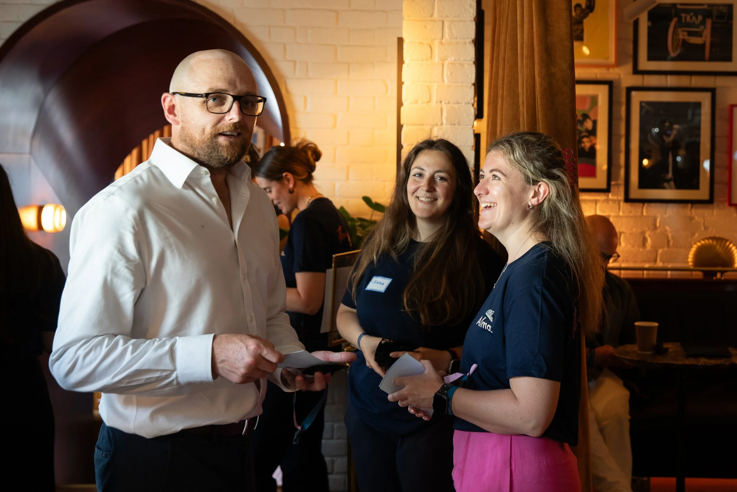 A group of four people in a restaurant, with three women wearing navy blue shirts with a logo and the man in a white shirt and glasses, engaging in conversation and smiling. The background shows a brick wall, framed pictures, and warm lighting.