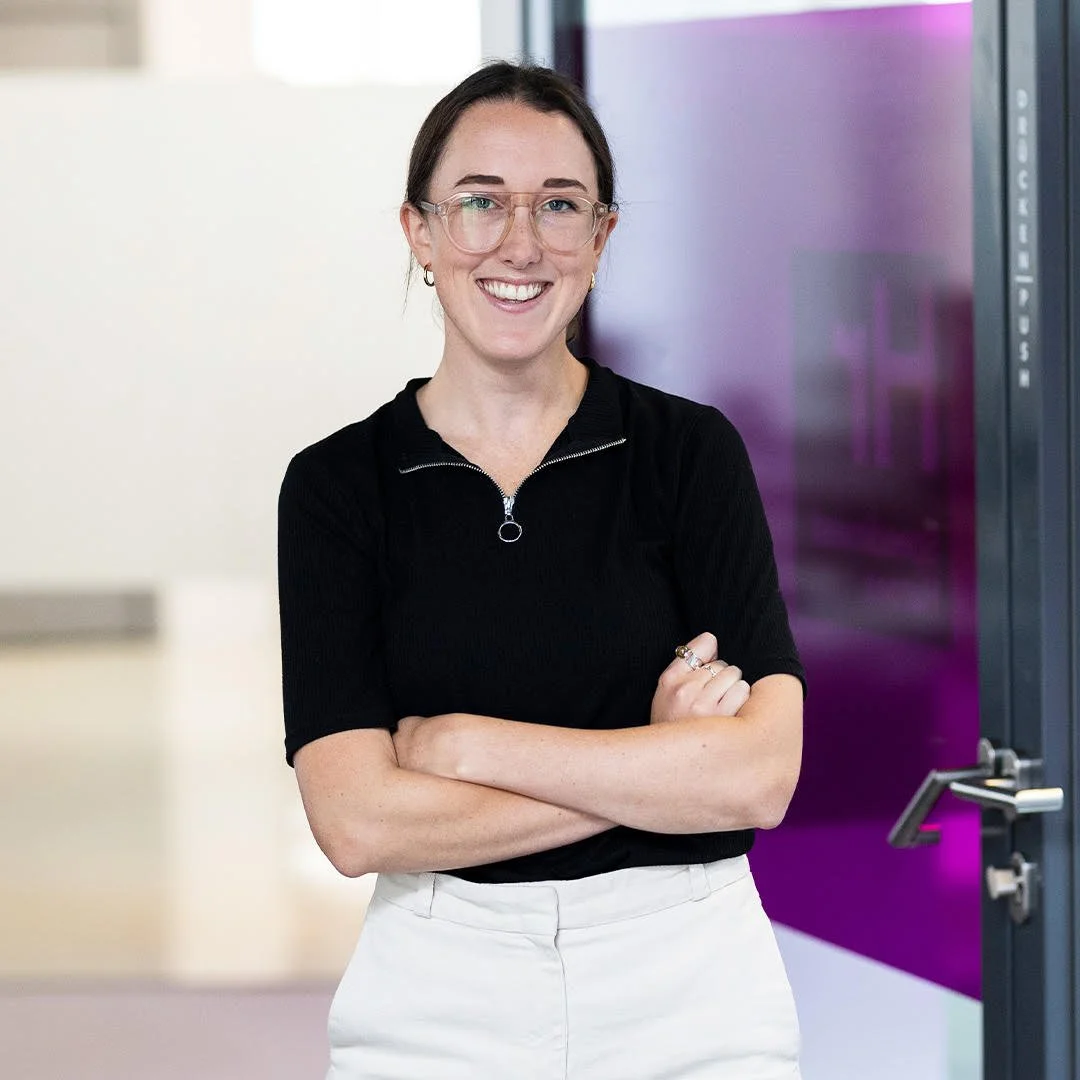 A woman with glasses smiling and crossing her arms, standing in front of a purple and gray wall.