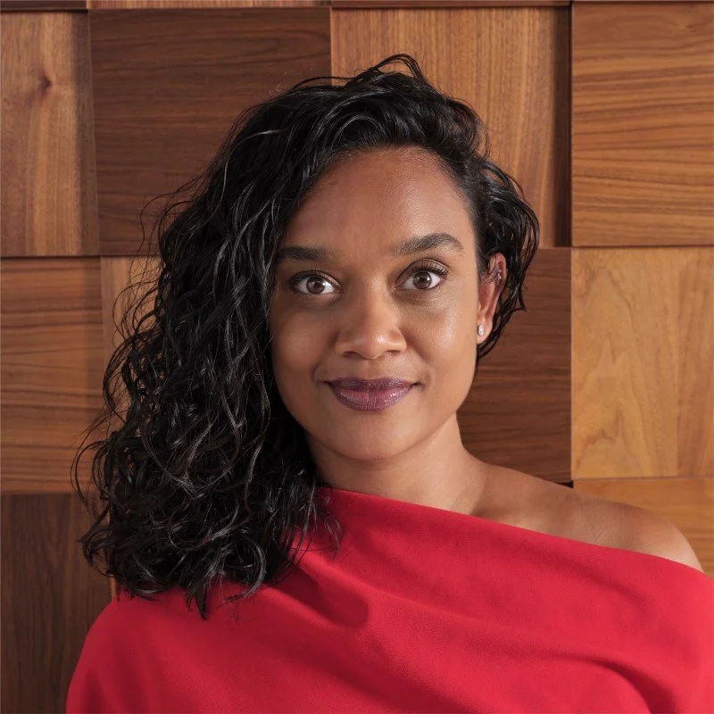 A woman with dark, curly hair wearing a red off-the-shoulder top, standing in front of a wooden paneled background.