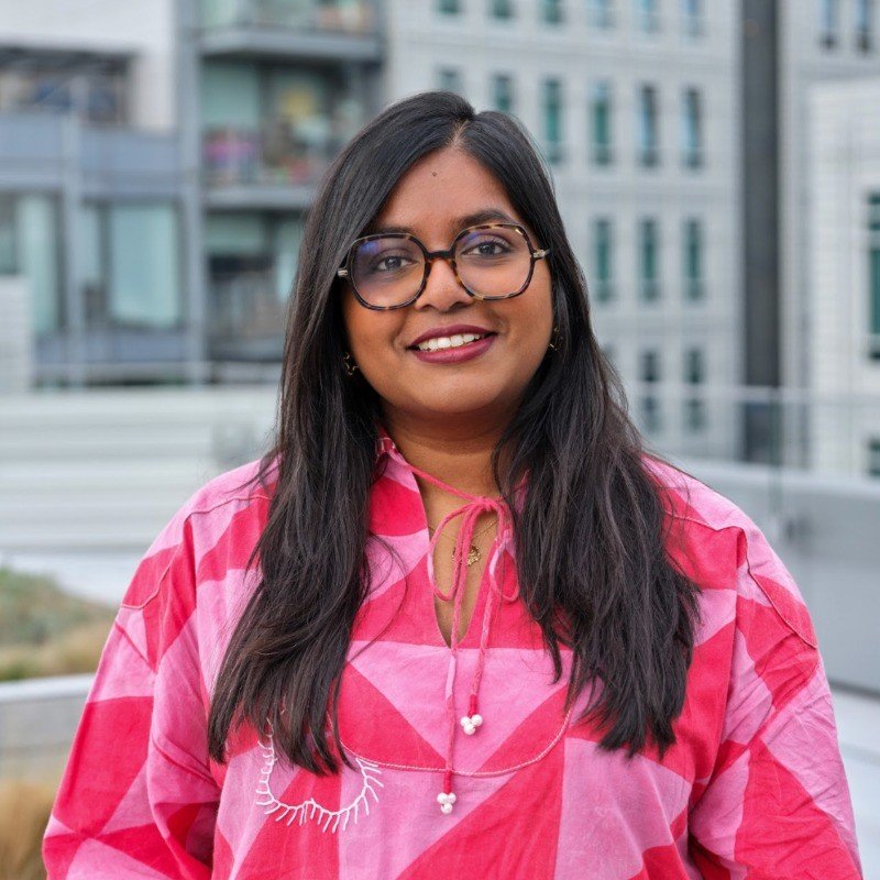 A woman with long dark hair and glasses, smiling, wearing a pink patterned shirt with a necklace, standing outdoors with a cityscape in the background.