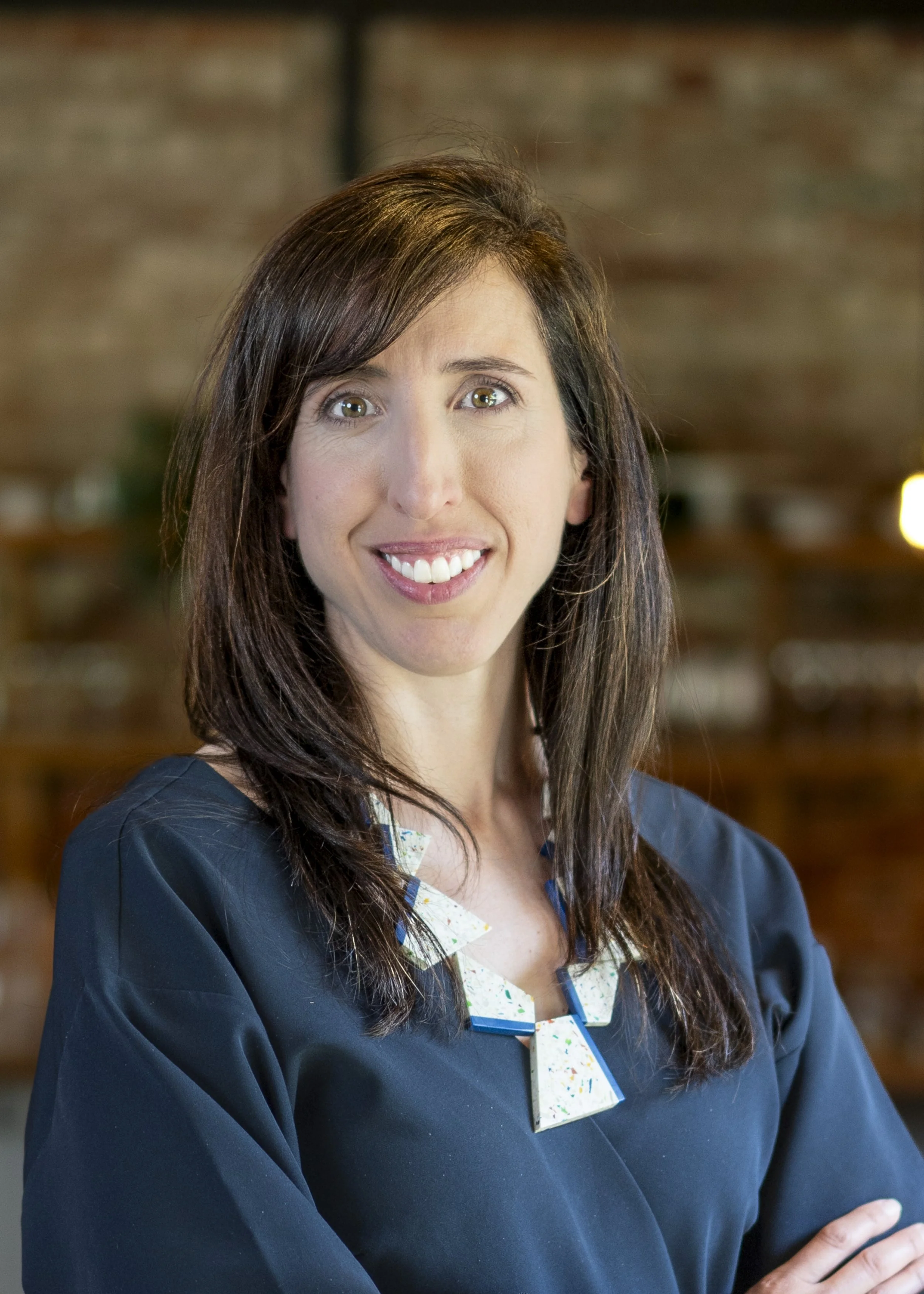 A woman with long brown hair and hazel eyes wears a dark blue academic gown and a white collared shirt with a colorful pattern. She is smiling and standing indoors with a brick wall background.