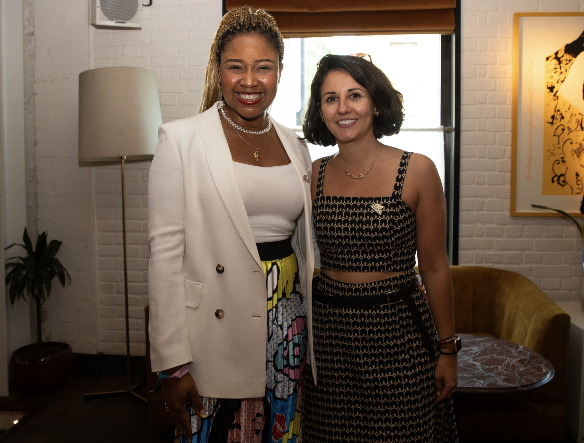 Two women standing side by side, smiling, inside a room with brick walls, a window, a floor lamp, a potted plant, and framed artwork on the wall.