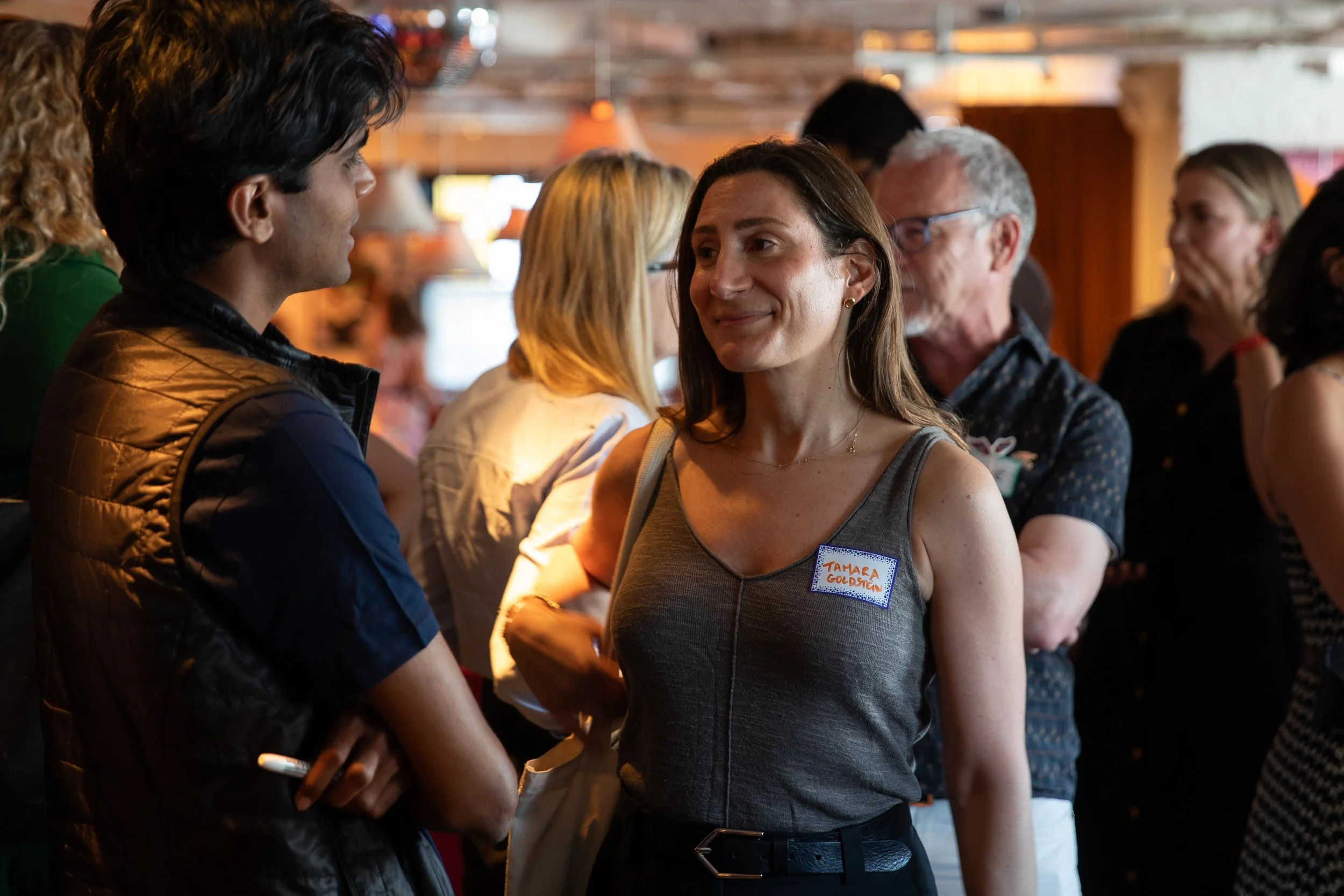 A group of people at a social gathering, including a woman with a name tag that reads 'TAMERA GOLDSTEIN', is engaged in conversation inside a warmly lit, rustic venue with wood decor.
