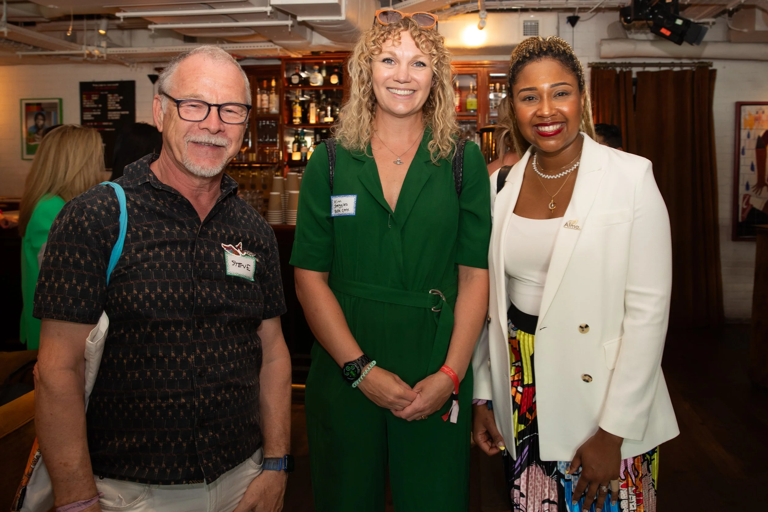 Three diverse people standing together in a bar or restaurant, smiling at the camera. The man on the left is wearing glasses, a patterned shirt, and has a name tag that says "Steve." The woman in the middle has curly blonde hair, sunglasses on her he