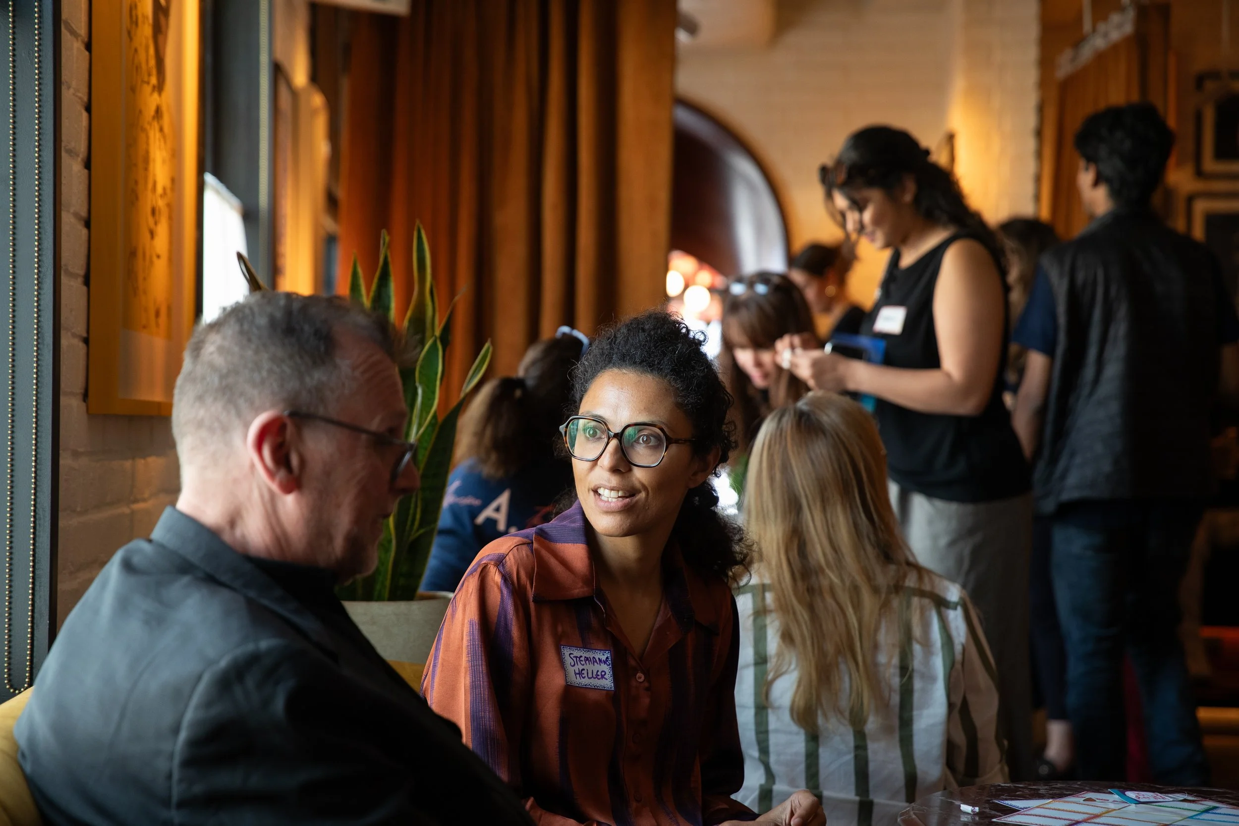 People gathered around a table at a social event, engaged in conversation, with warm lighting and wooden decor in the background.