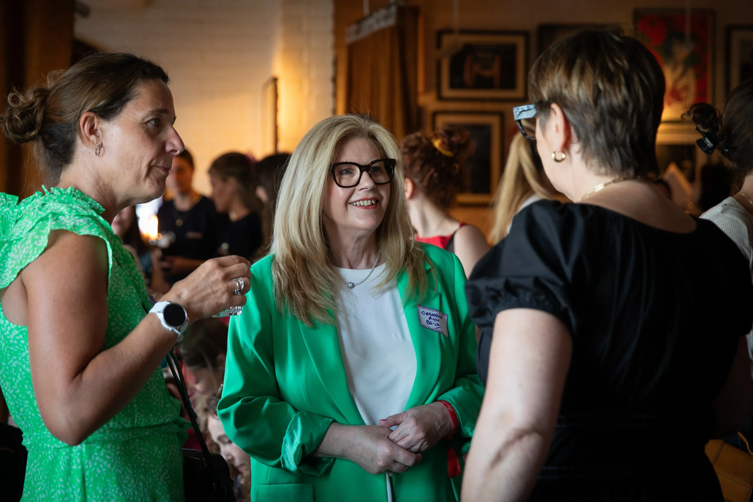 Three women engaged in conversation at a social gathering indoors, with a woman in a bright green jacket smiling in the center, wearing glasses, surrounded by two other women, one in a green dress and the other in a black top.