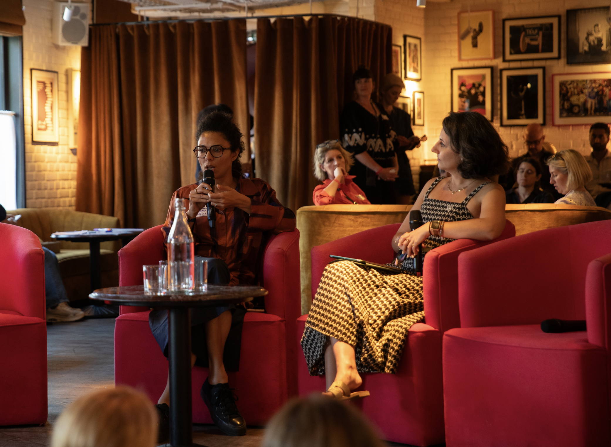 People sitting on red chairs in a cozy room with artwork on the walls and two women standing behind a curtain in the background.