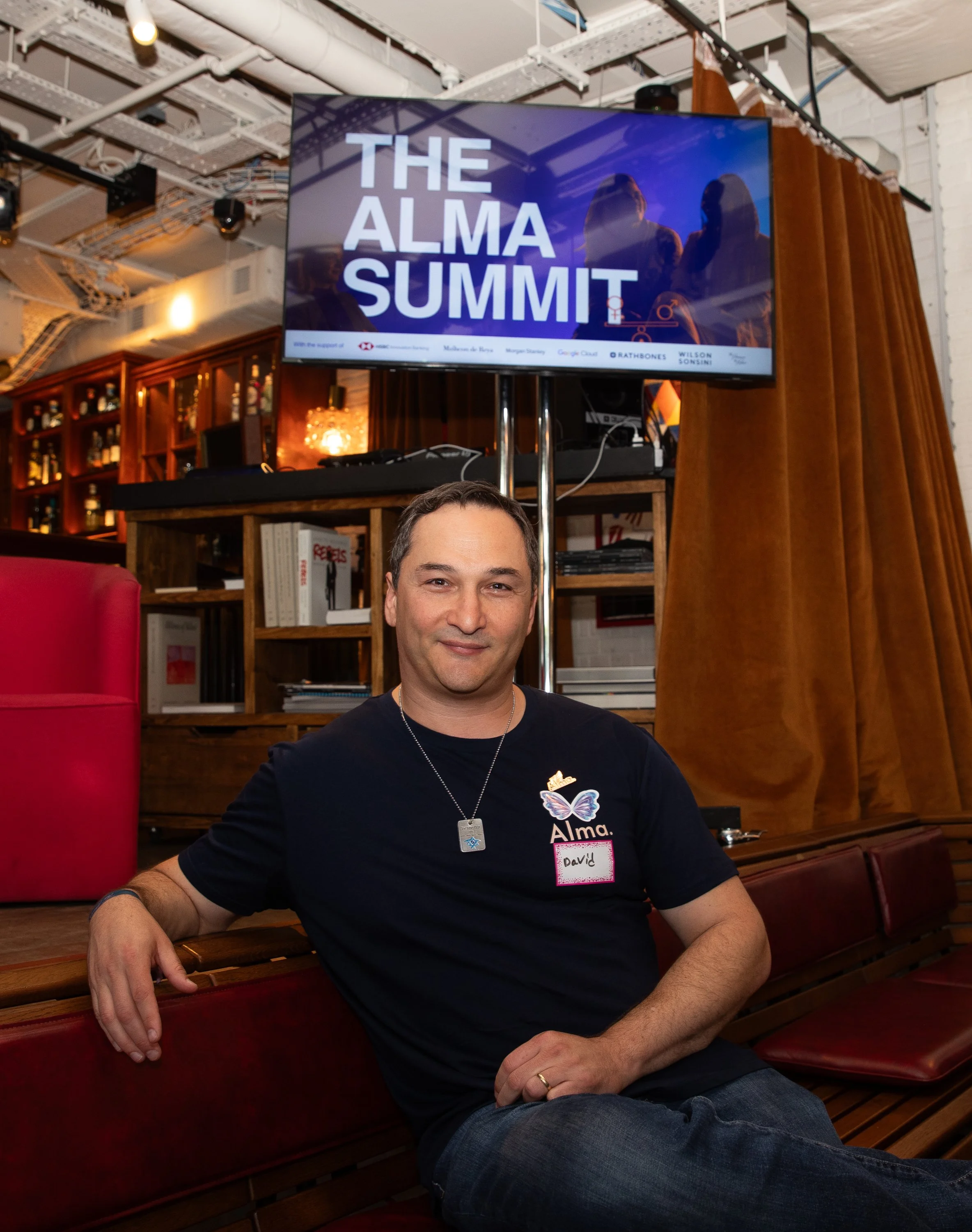 A man with short dark hair and wearing a black T-shirt with a name tag that says 'David' and a butterfly logo sitting on a red bench in front of a bookshelf and a TV screen that reads 'The Alma Summit' in a room with exposed ceiling pipes.