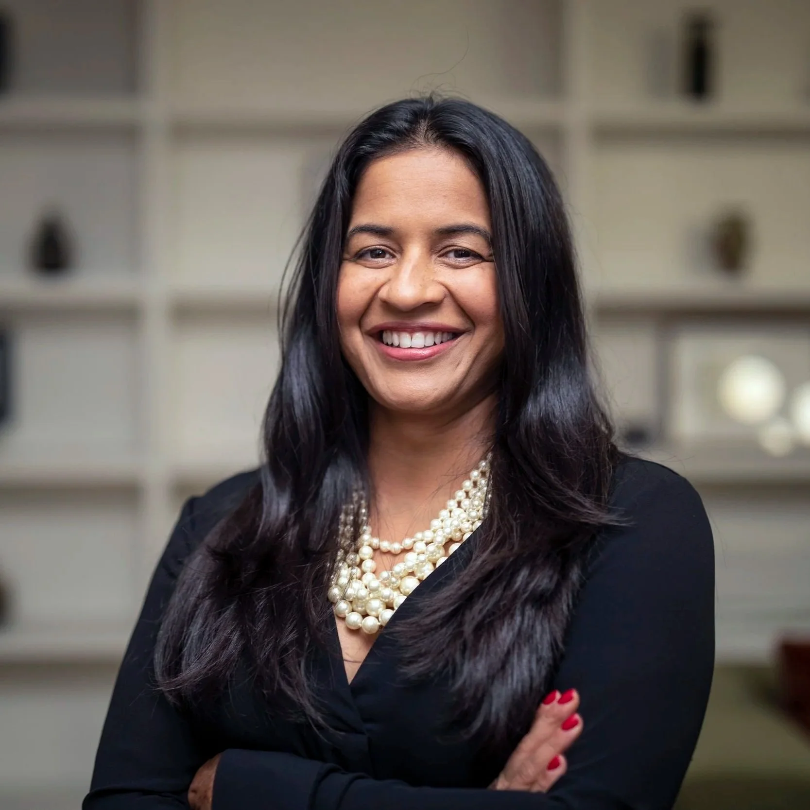 A woman with long dark hair wearing a black blazer and layered pearl necklaces, smiling confidently, standing in front of a neutral background with shelving.