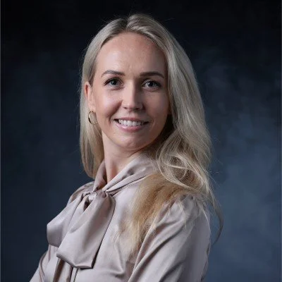 A young blonde woman with long hair smiling against a dark background.