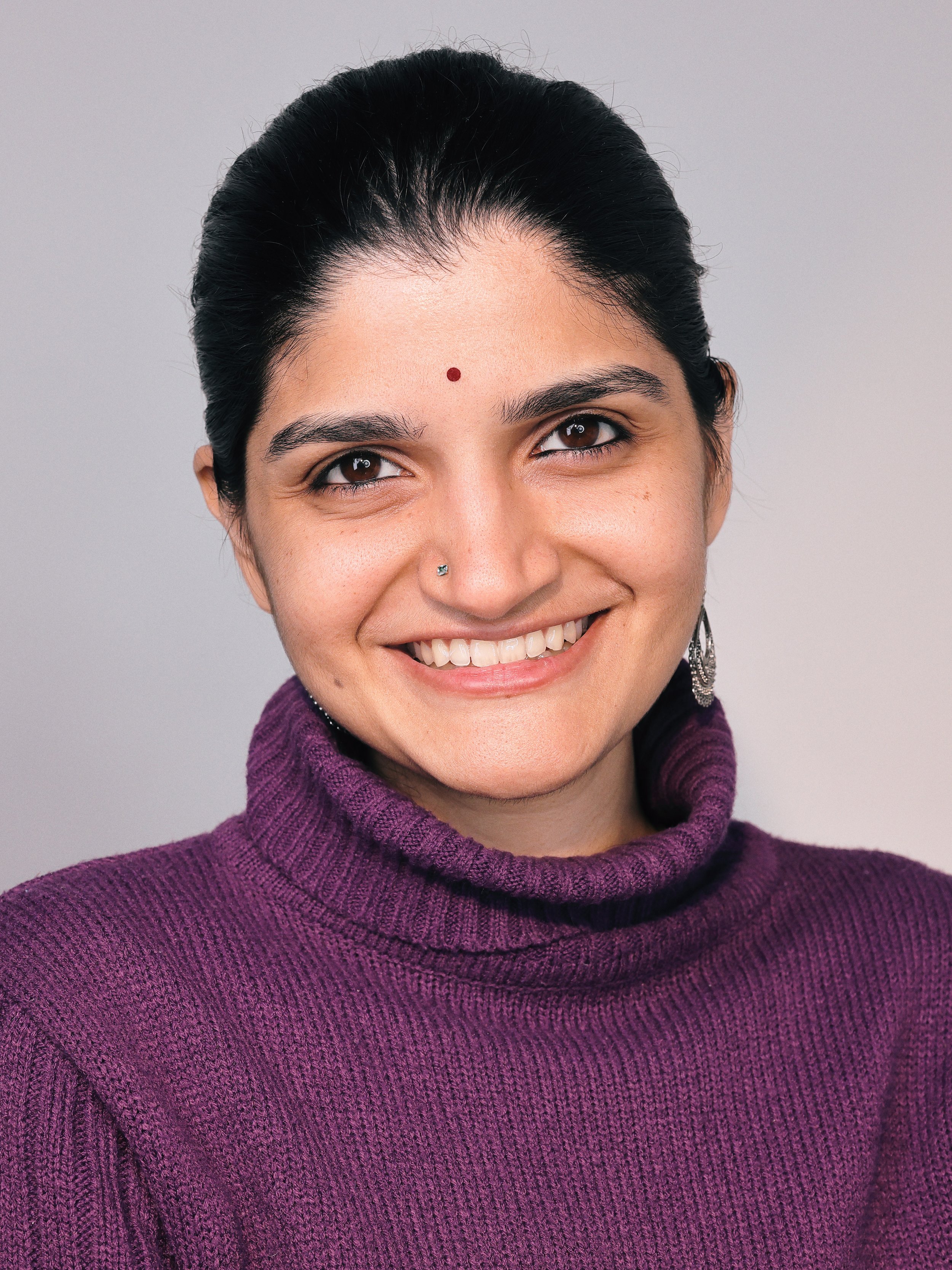 Close-up of a smiling woman with black hair, wearing earrings, a purple turtleneck sweater, and a small red bindi on her forehead against a plain gray background.