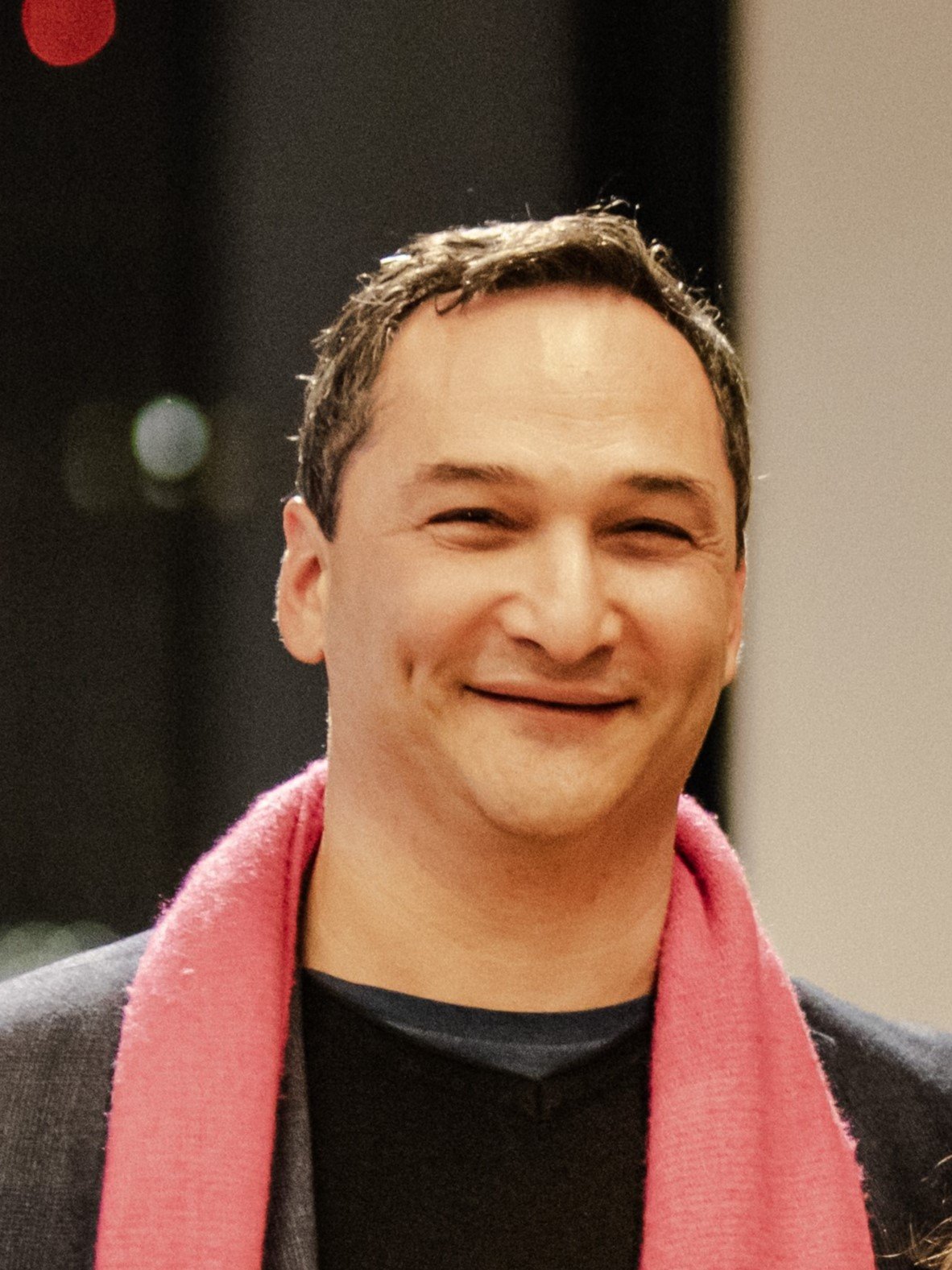 A man smiling with closed eyes, wearing a dark jacket and a red scarf.