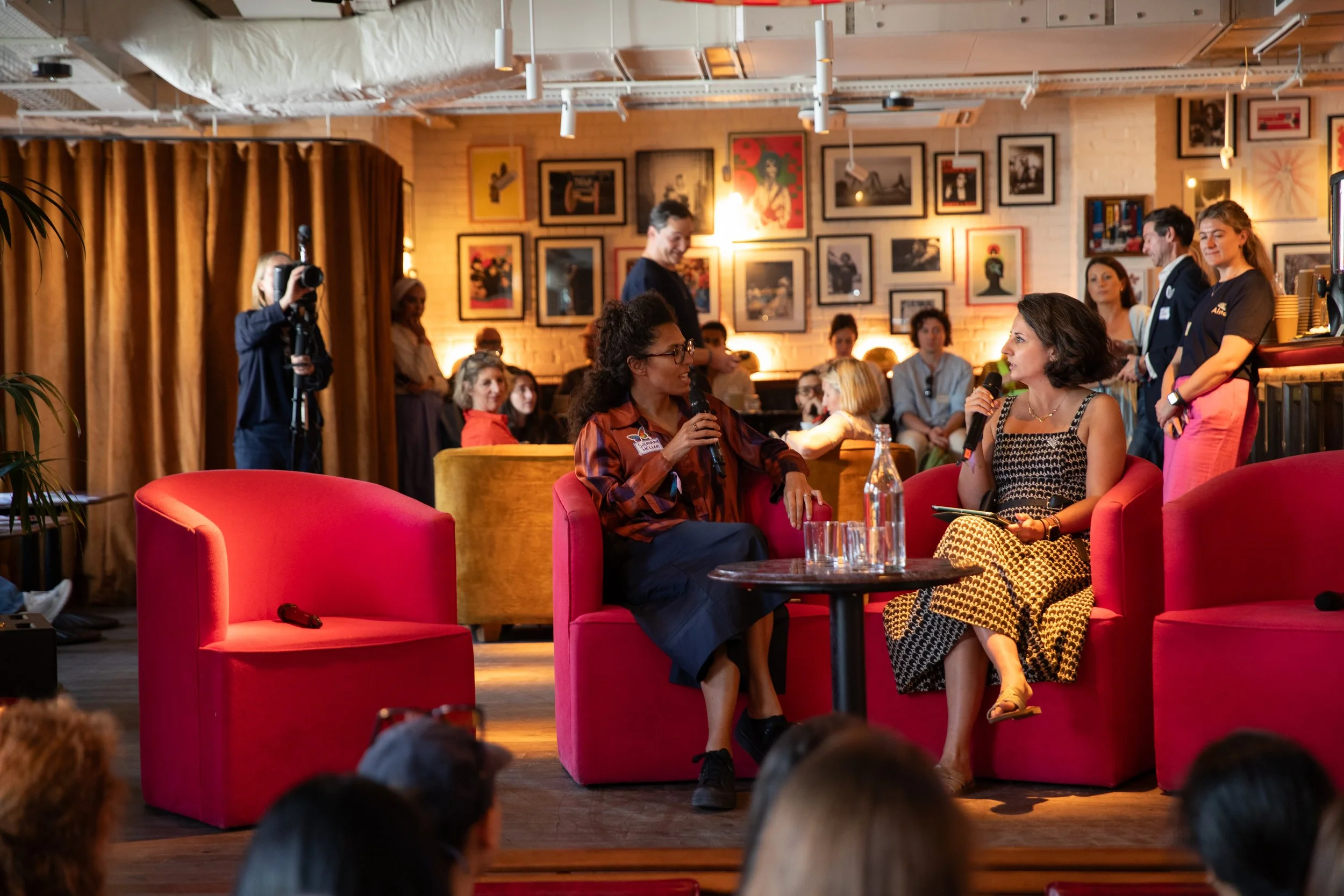 A group of people at an indoor event, with two women sitting and speaking into microphones while others listen. The setting is a cozy room with framed artwork on the walls and warm lighting.