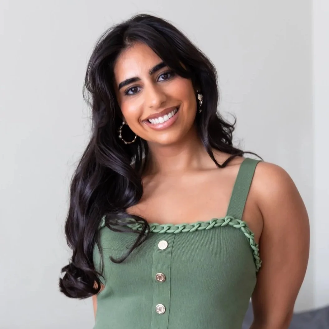 A woman with long dark hair, wearing a green sleeveless top with decorative buttons and braided trim along the neckline, smiling at the camera.