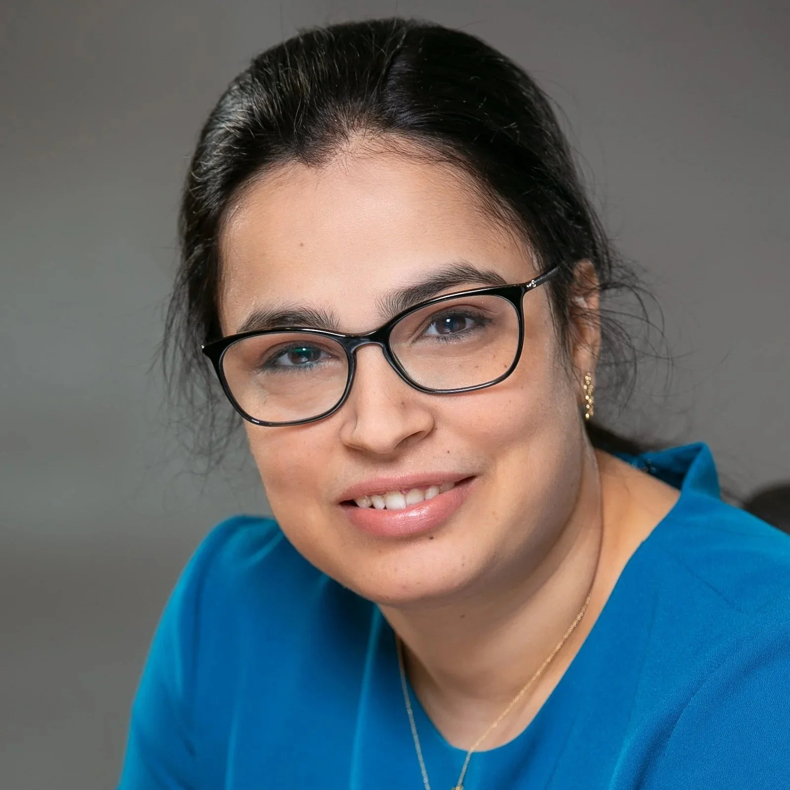 A woman with dark hair, glasses, and gold earrings, smiling, wearing a blue top, against a grey background.
