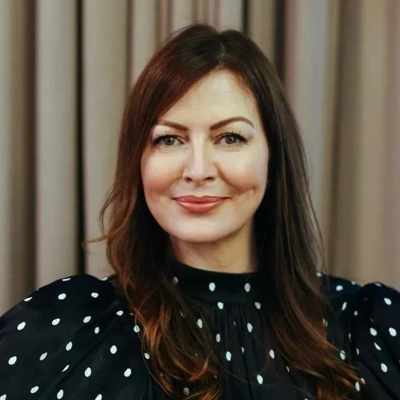 A woman with shoulder-length brown hair, smiling, wearing a black dress with white polka dots, standing in front of beige curtains.