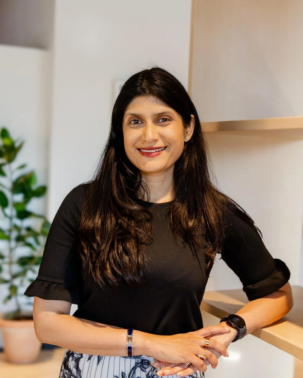 A woman with long dark hair smiling and standing indoors. She is wearing a black top and a black watch, with a bracelet and rings on her hands. There is a plant and a wooden surface in the background.