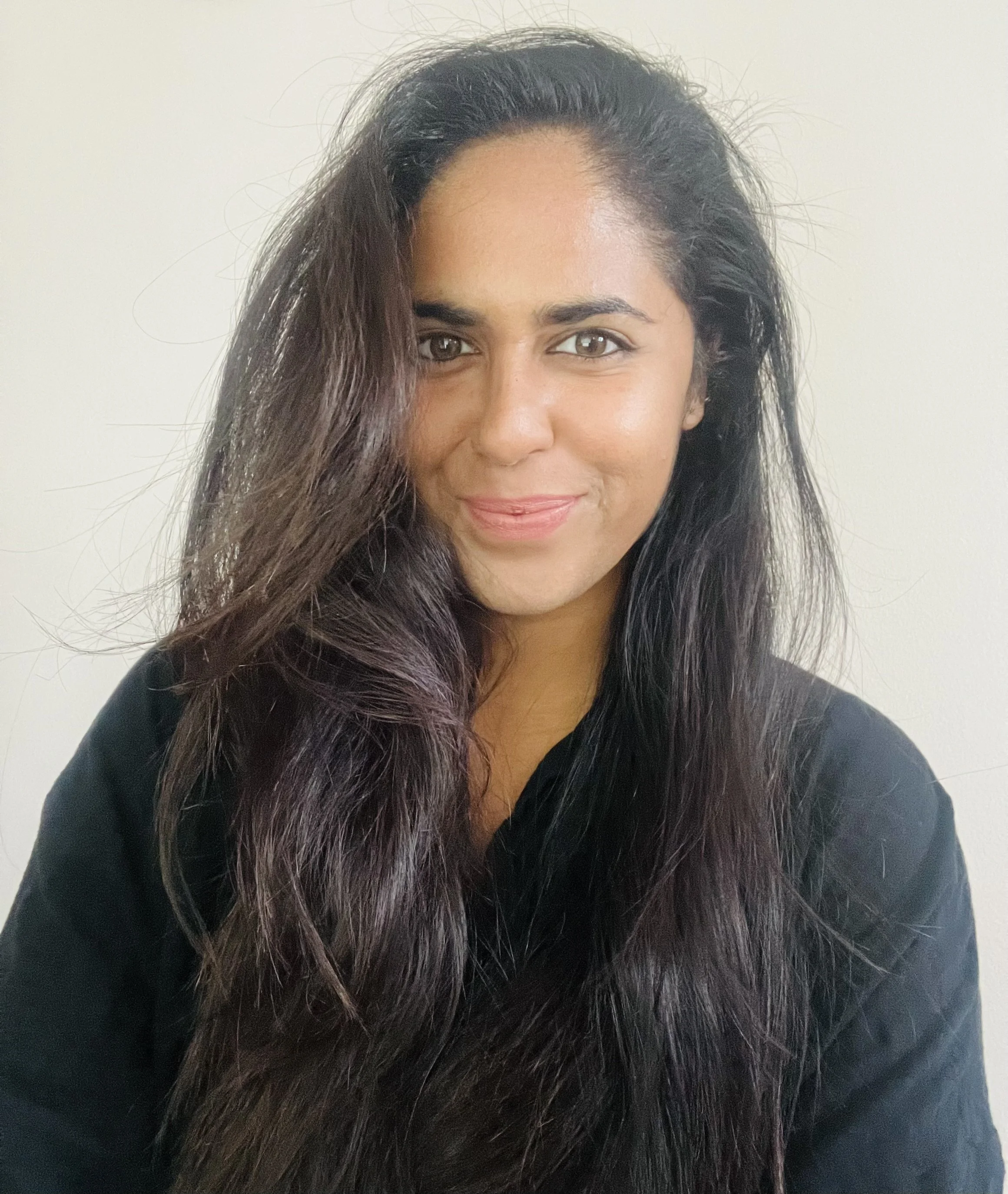 Close-up of a woman with long dark wavy hair, smiling softly, wearing a black top, against a plain light background.