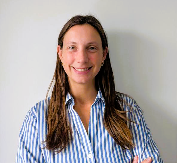 A woman with long brown hair wearing a blue and white striped button-up shirt, smiling and standing with her arms crossed in front of a plain light-colored wall.