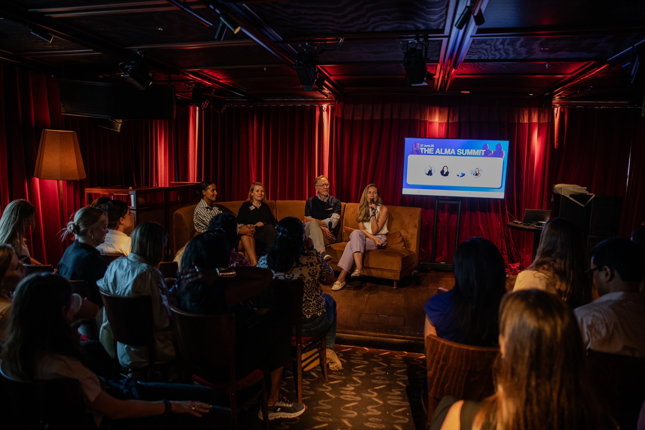 Panel discussion at an event called The Alma Summit, with four speakers sitting on a brown couch on stage, a blue screen behind them displaying the event's name and date, red curtains surround the stage, and a large audience seated in front.