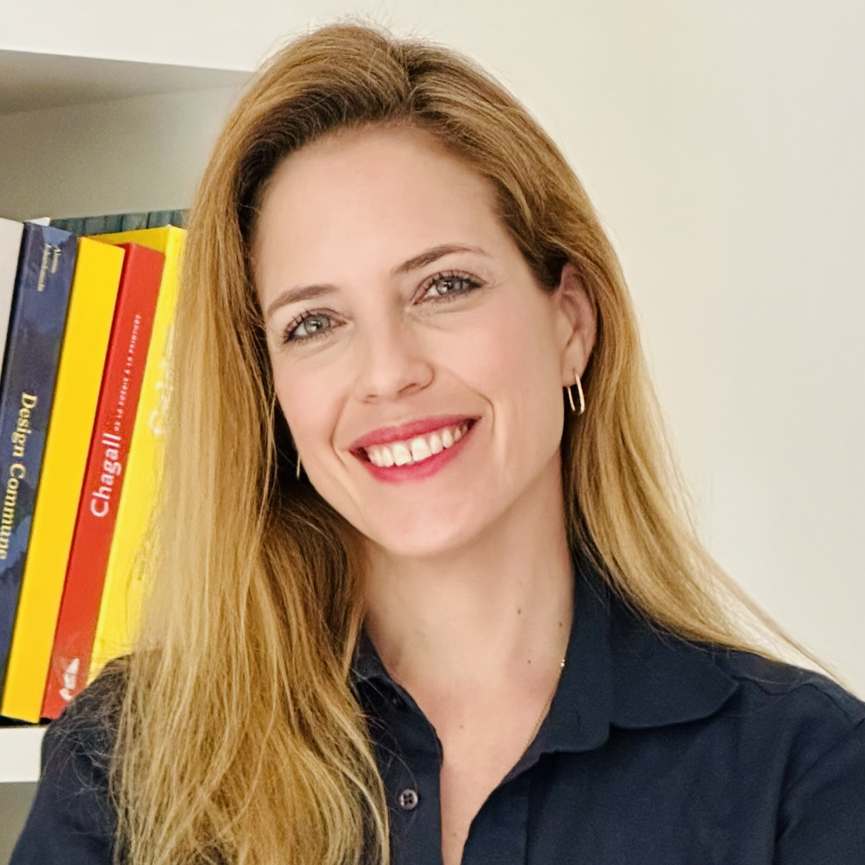 A woman with long red hair smiling and wearing a black shirt, standing in front of a white wall with books on a shelf.