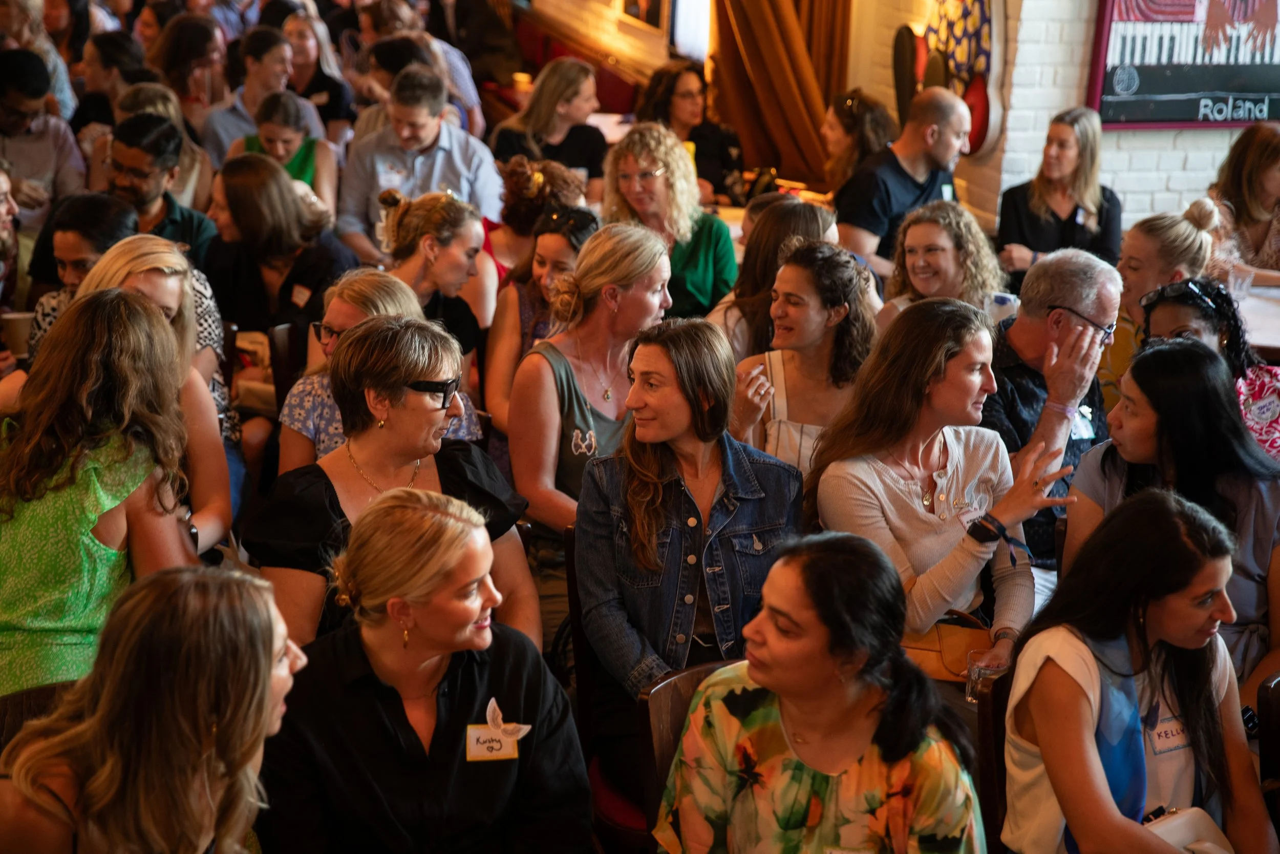 A large group of people gathered in a cozy indoor setting, engaging in conversations. The room has wood-paneled walls and a chalkboard with the word 'Roland' written on it.