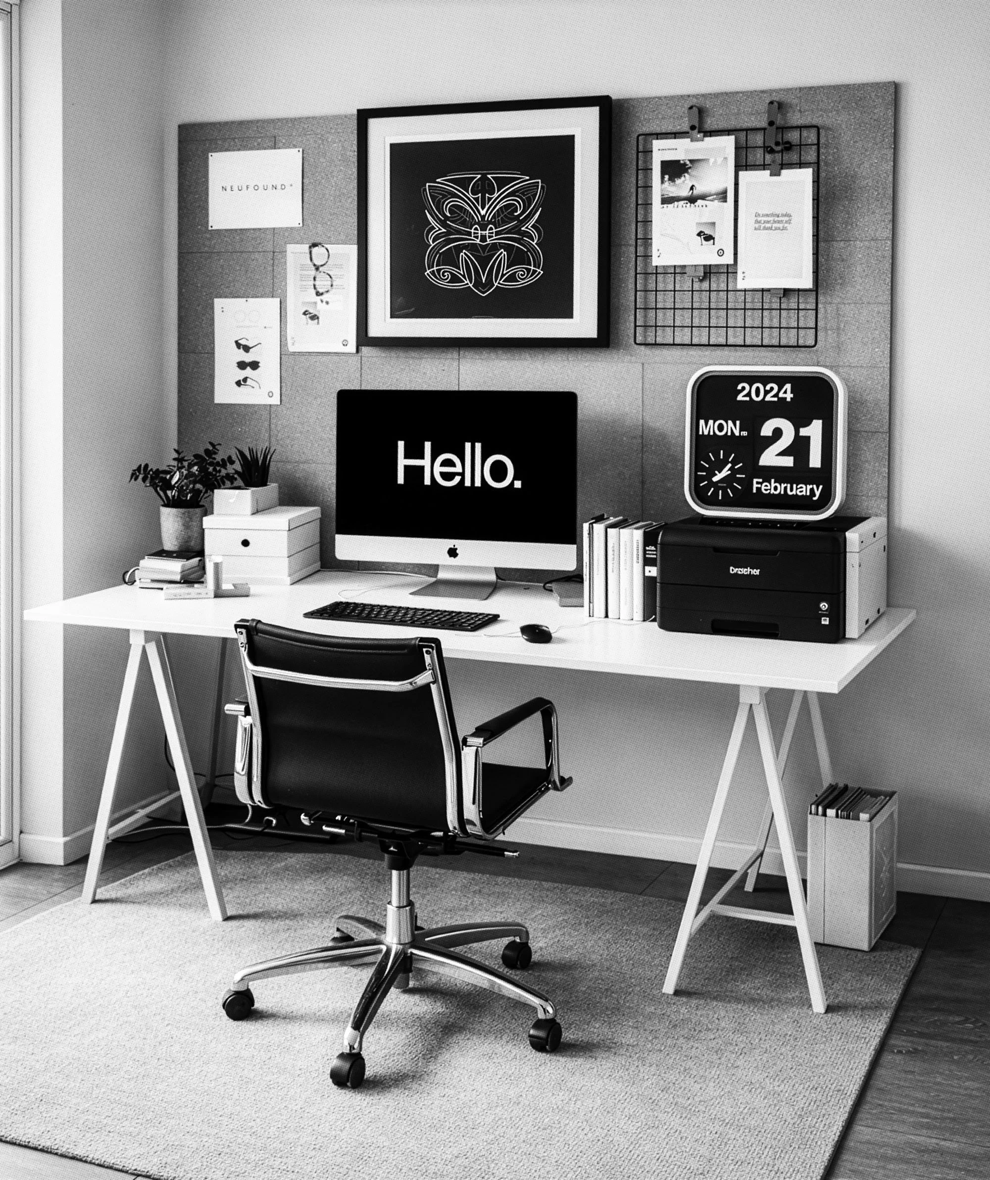 Black and white photo of a modern office workspace with a white desk, an adjustable chair, a desktop computer displaying the word 'Hello.', a desk calendar showing February 21, 2024, and various decorative items on the wall, including framed art and a grid with papers.