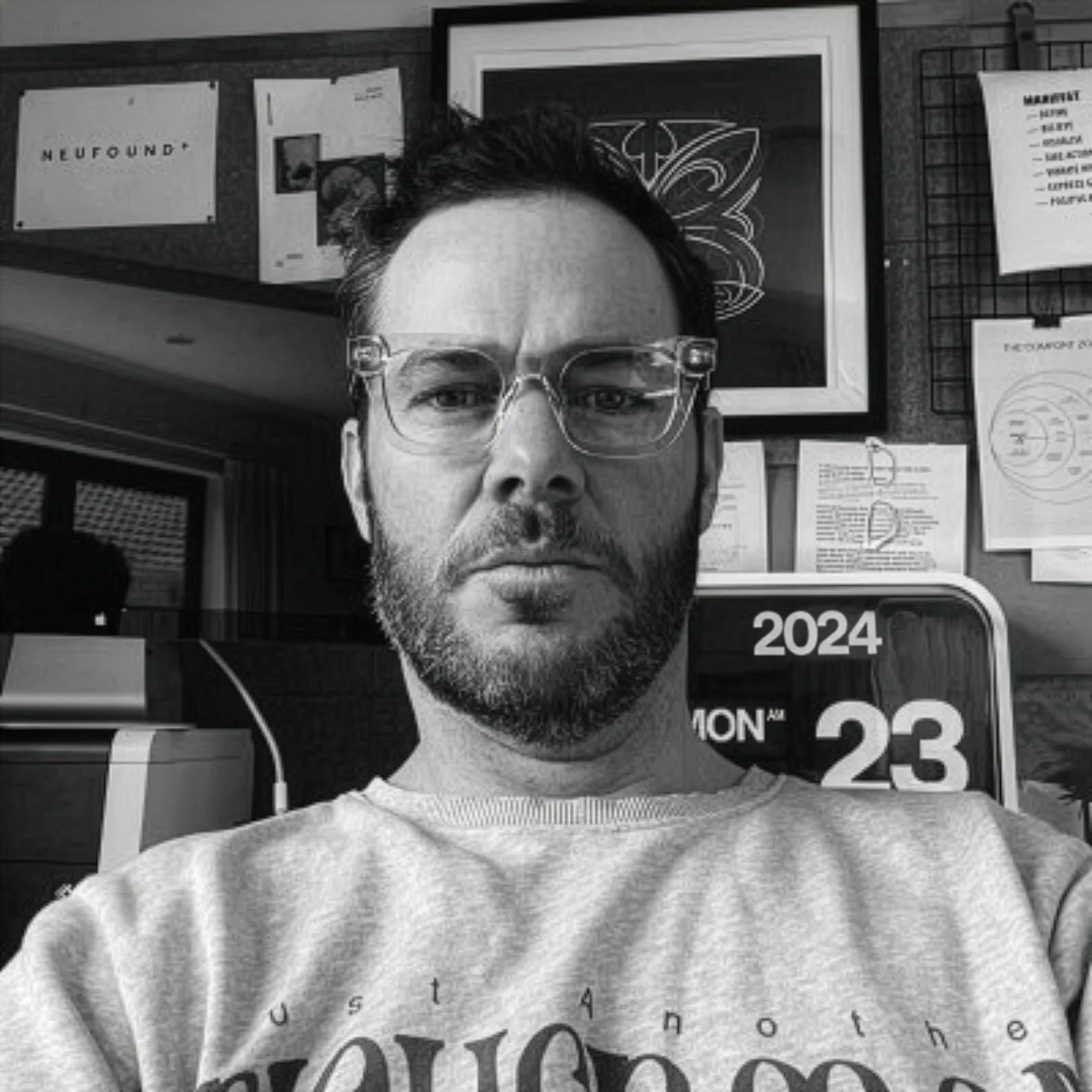 A man with glasses and a beard taking a selfie indoors, with a background that includes framed artwork, papers, and a digital calendar showing April 23, 2024.