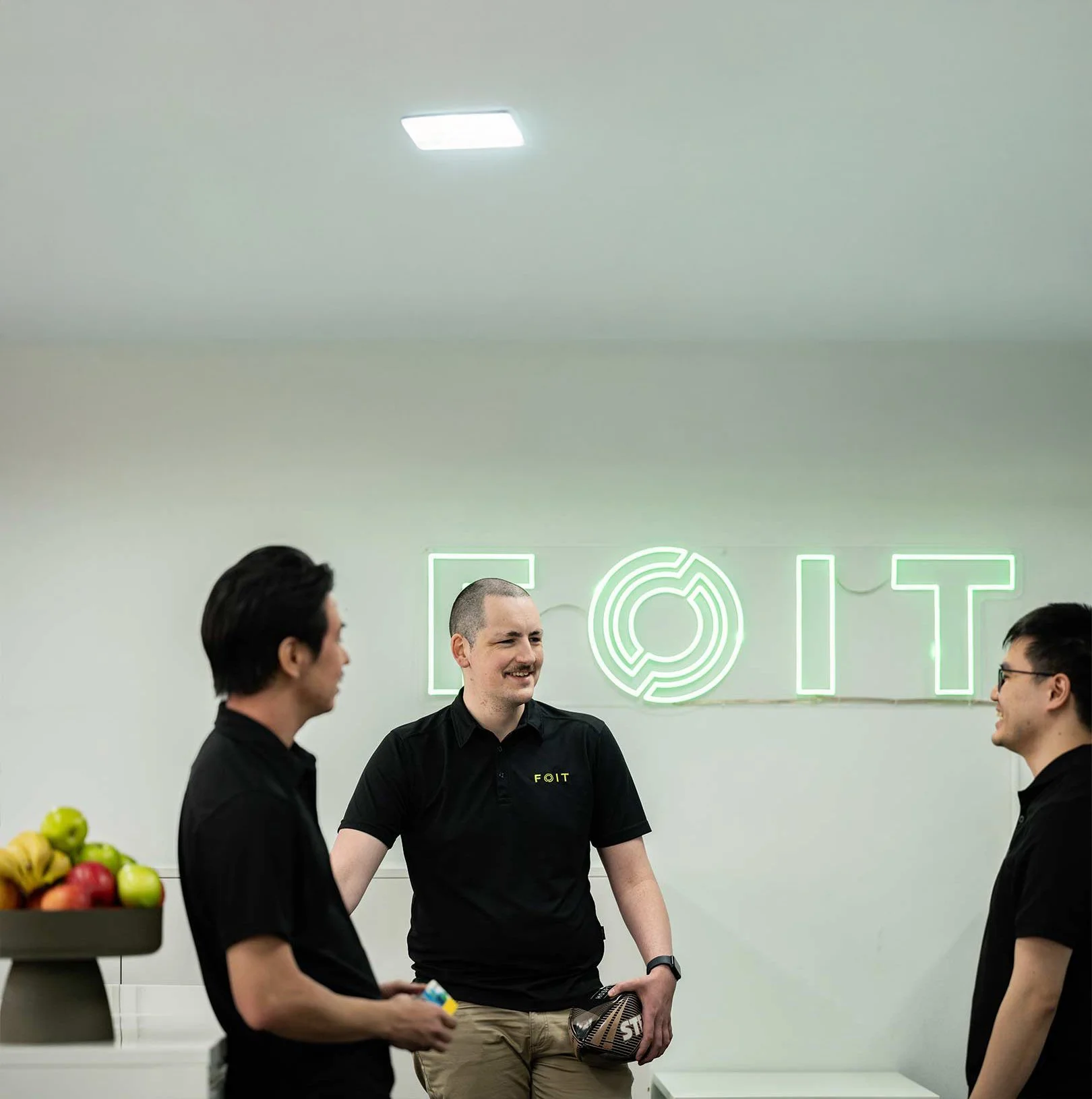 Three men in black shirts standing and talking in an office with a neon sign that reads 'FOIT' on the wall.