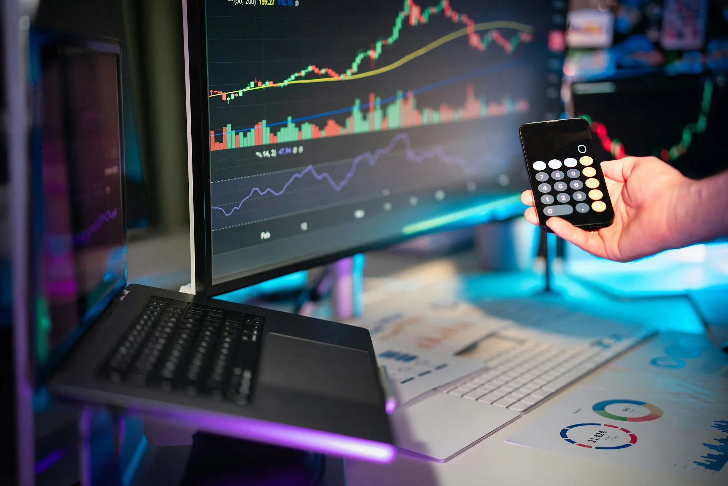 A person holding a smartphone with a calculator app in front of a computer monitor displaying stock market charts and graphs in a workspace with papers, a keyboard, and a laptop.