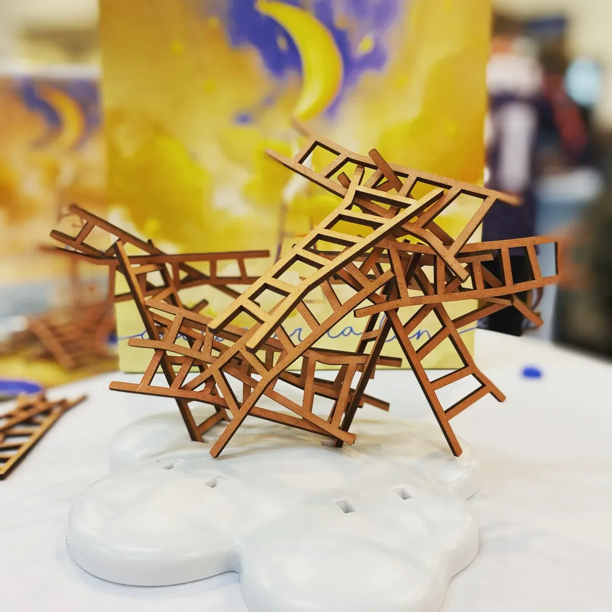 A small, intricate wooden sculpture of several overlapping ladders placed on a white, cloud-shaped base. In the background, a yellow container with a crescent moon and star design is visible.