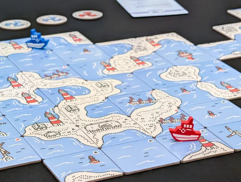 A board game featuring an illustrated map of an island with lighthouses and water, with small ship game pieces, including a red ship and a blue ship, on a black table. There are also circular game tokens at the top of the image.