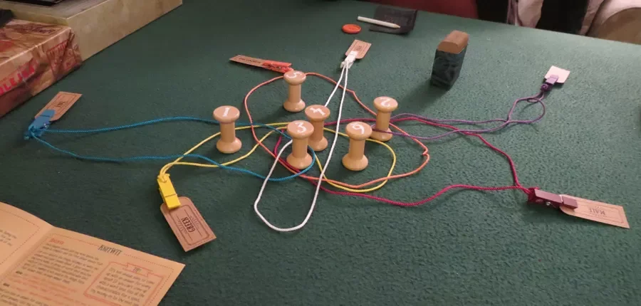 A tabletop board game setup with wooden pegs, colored strings, and various paper and plastic components, resembling an educational or puzzle game.