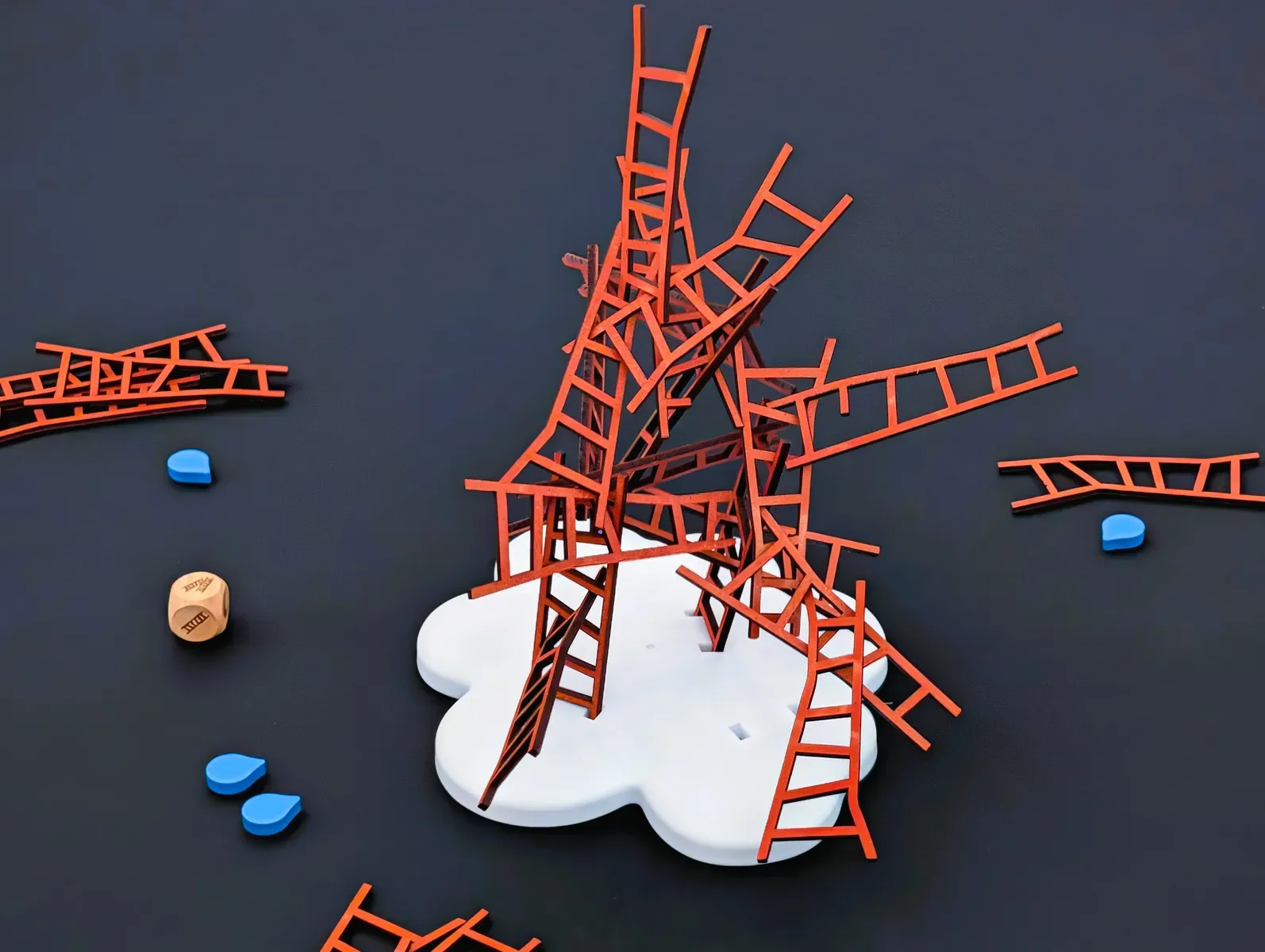 A tabletop game with orange ladder pieces assembled in a pile on a white base, with scattered blue and beige game tokens around.