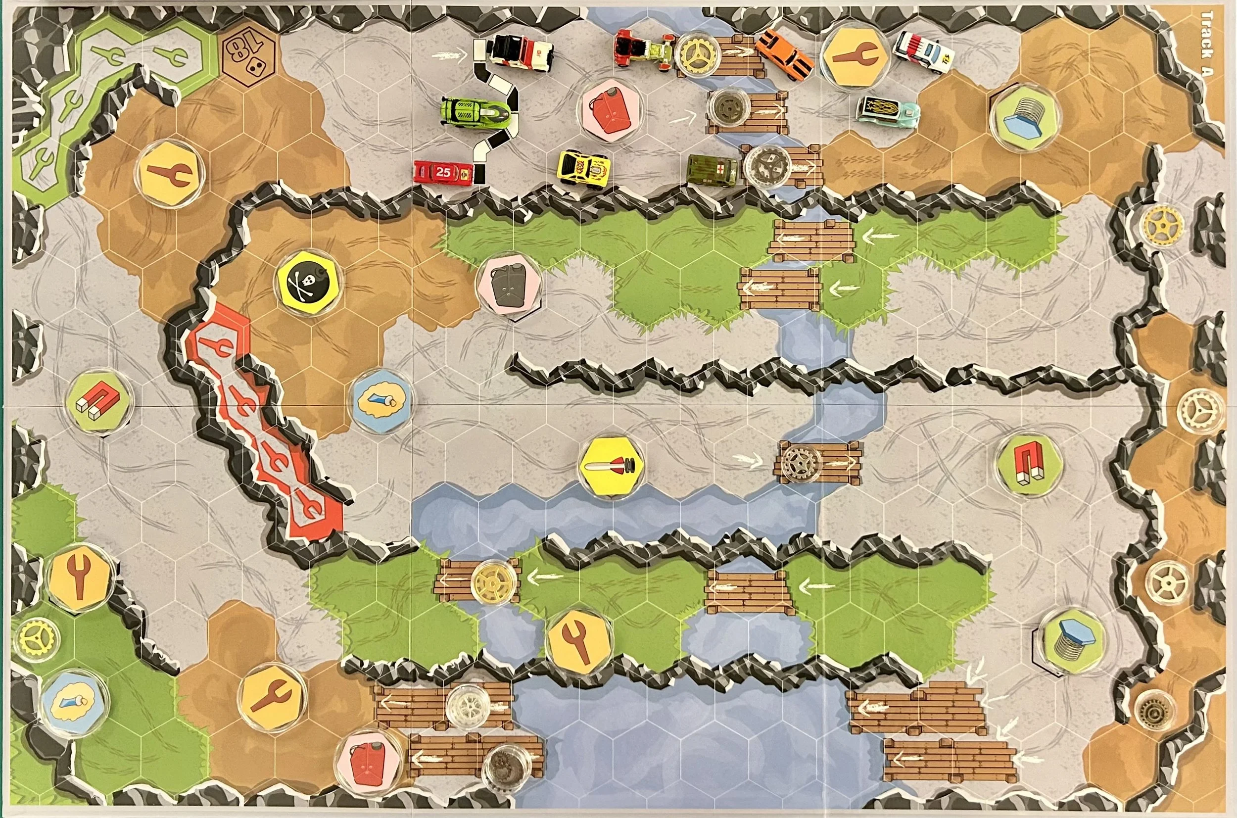 A tabletop board game with a hexagonal grid, featuring roads, rivers, bridges, resource tokens, and multiple toy vehicles including cars and trucks.