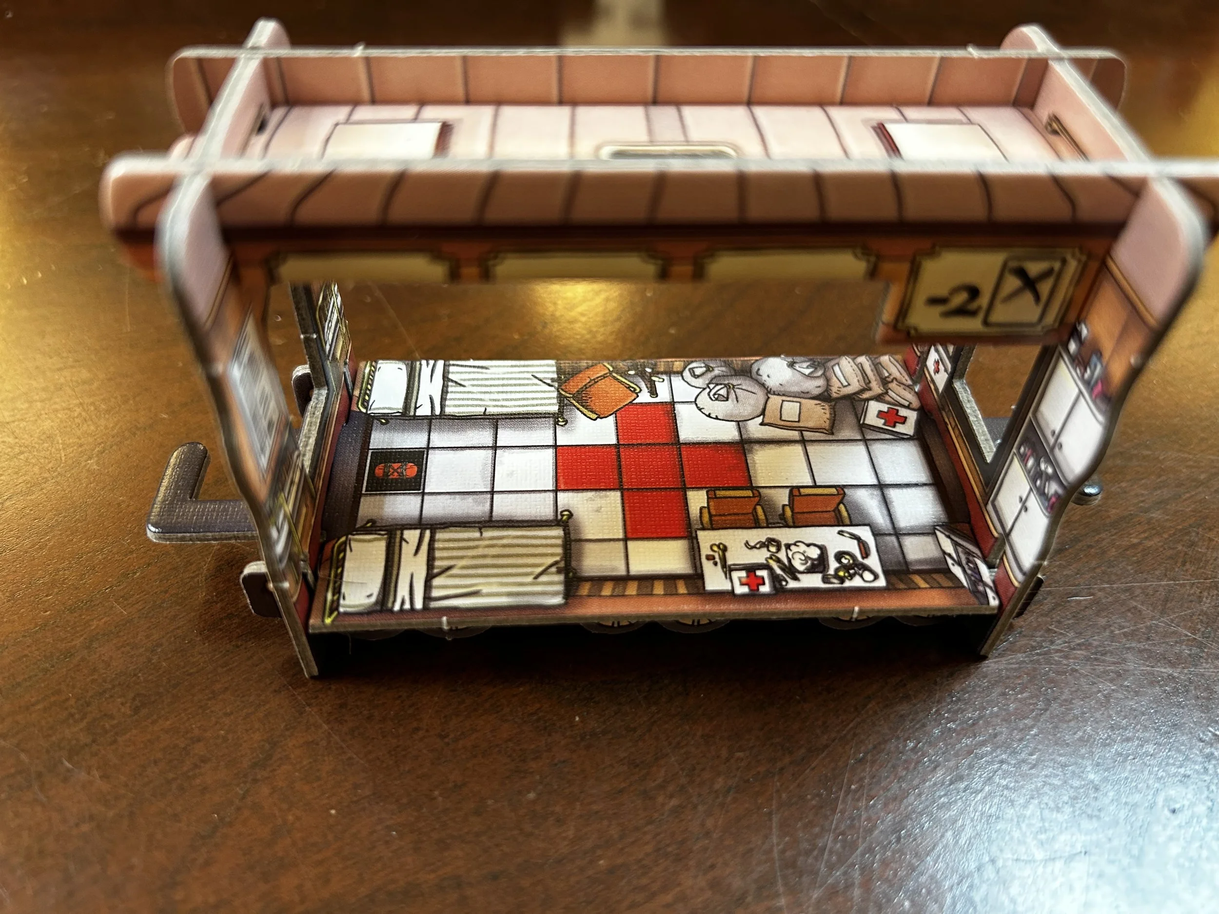 A miniature paper craft model of a hospital room with beds, medical supplies, and furniture, viewed from an angle overhead on a wooden surface.