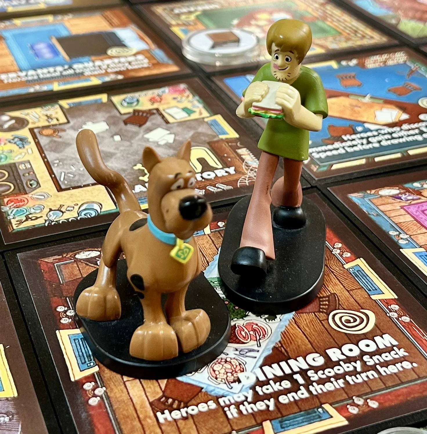Scooby-Doo and Shaggy figurines on the game board of Scooby-Doo Board Game, with colorful game tiles and instructions visible in the background.