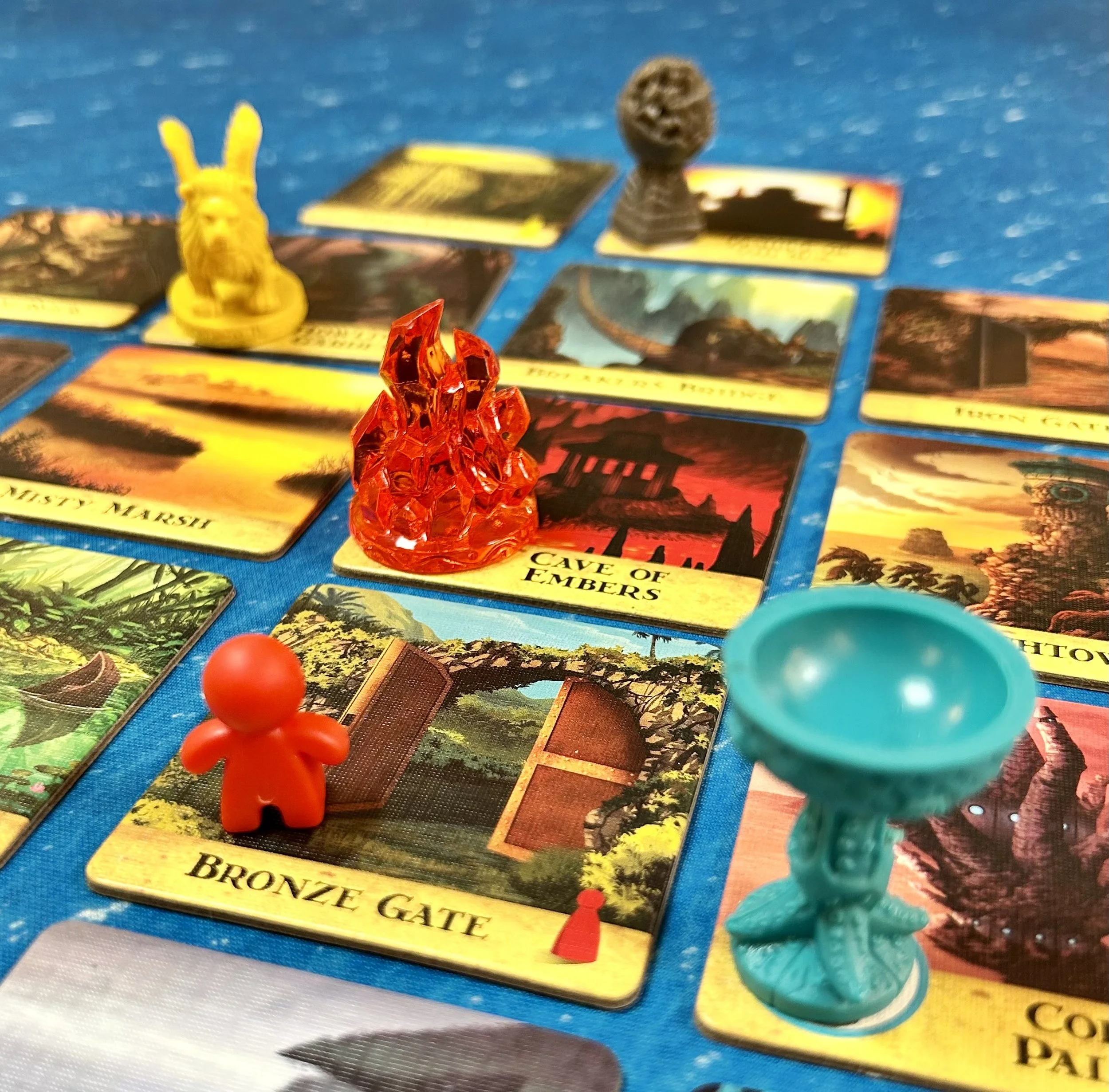 Board game with colorful illustrated cards, red, yellow, and blue game pieces, and miniature objects resembling a red fire, a yellow rabbit, and a gray tree, on a blue surface.