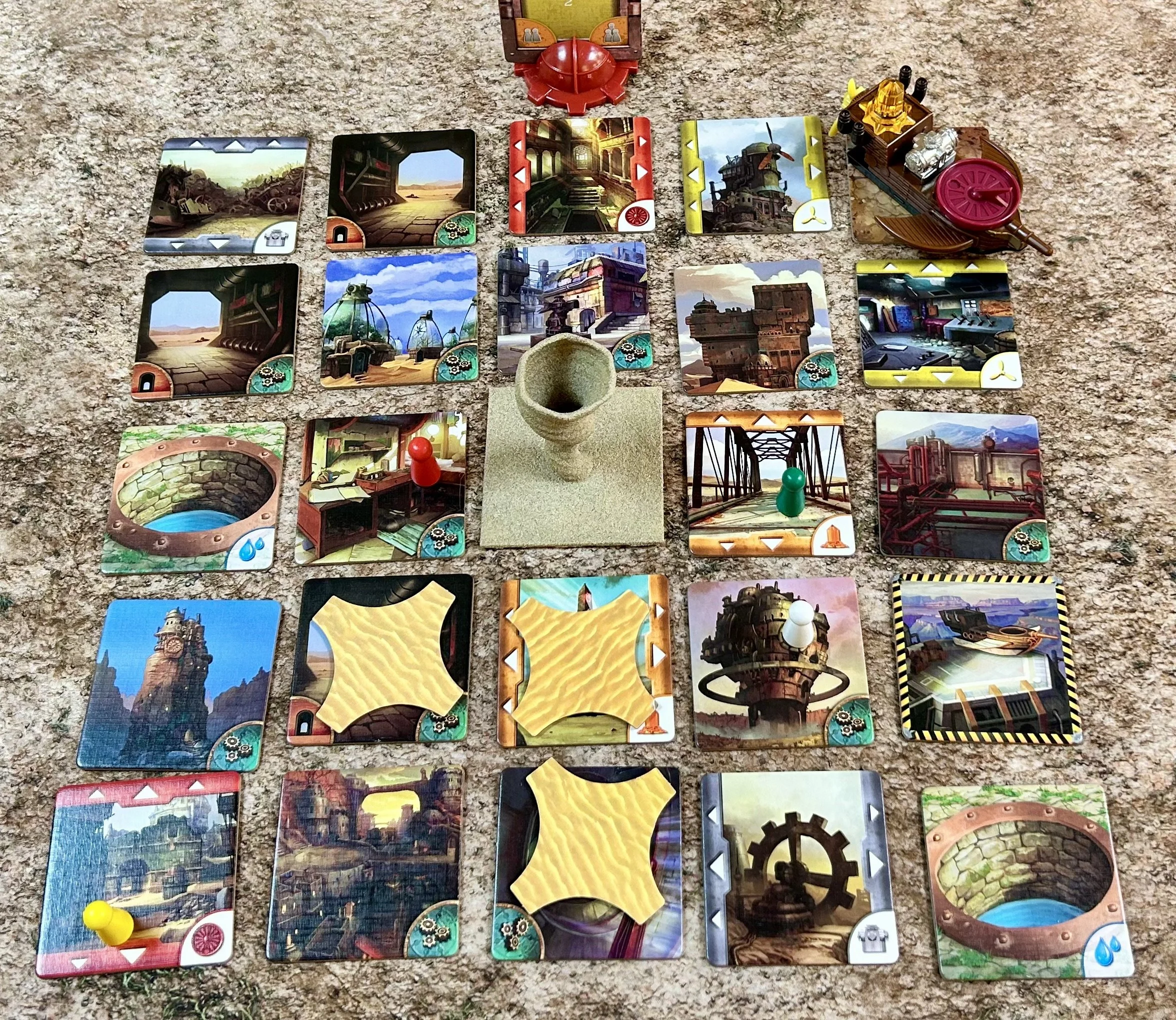 A tabletop game setup with various colorful tiles, tokens, and a boat. Some tiles depict industrial, city, and infrastructure scenes. The game pieces include a red pawn, a white pawn, a yellow pawn, and a boat, placed on a textured surface.